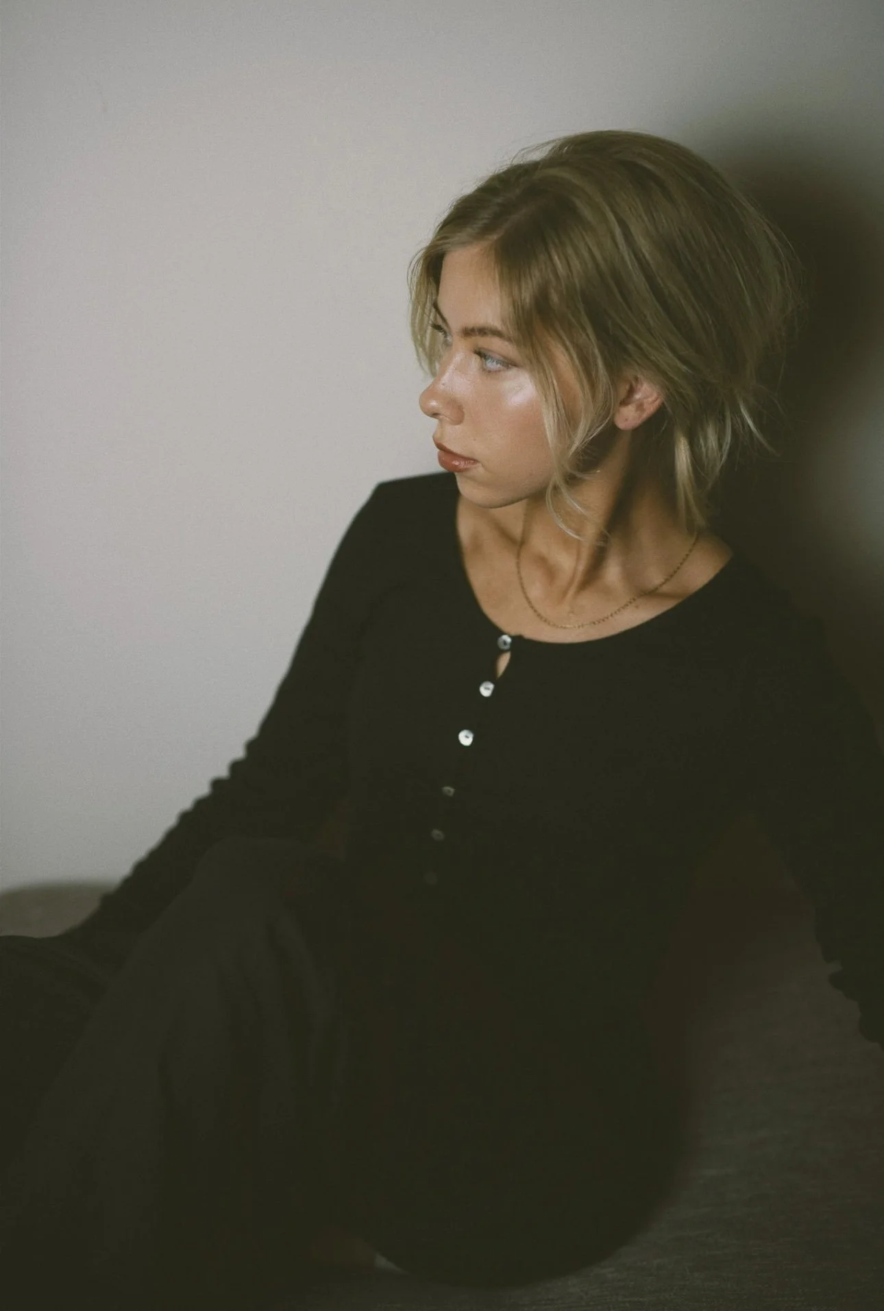 A woman with short blonde hair and a black top sitting against a plain white wall.