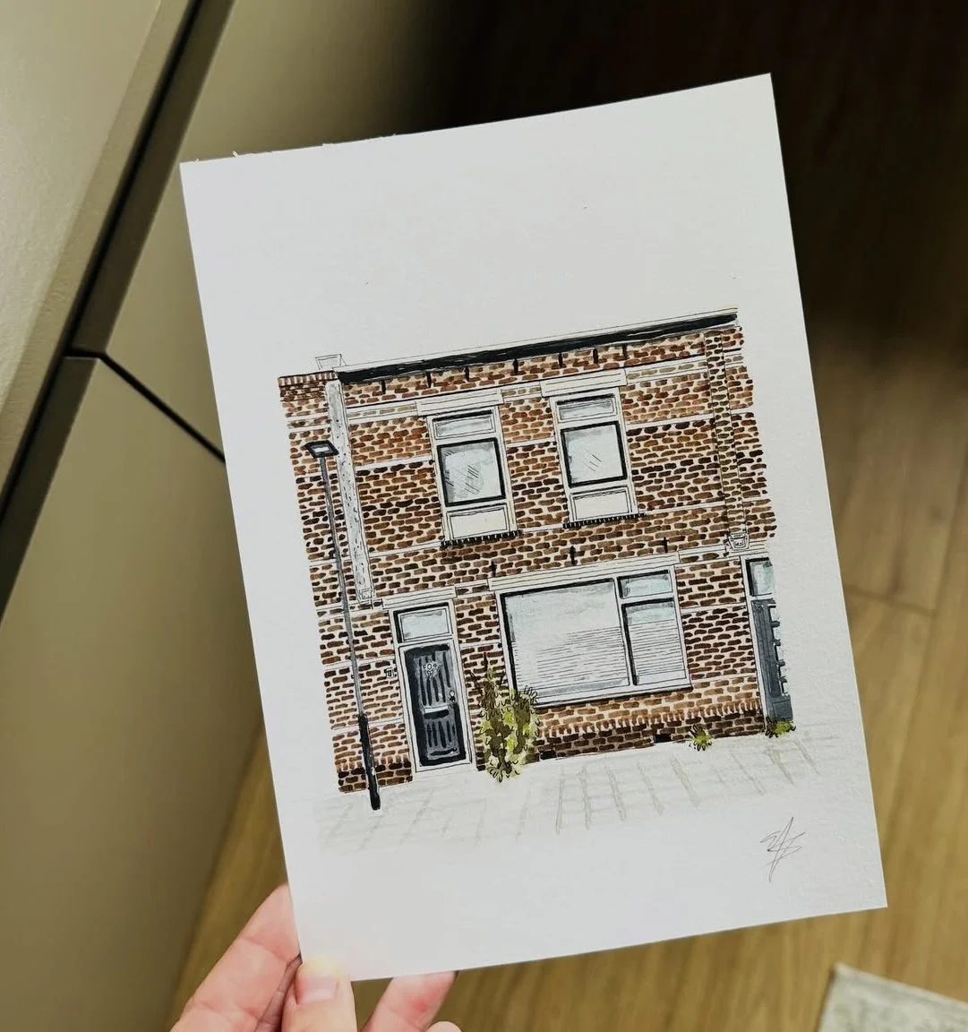 A hand holding an illustration of a brick building facade with two floors, windows, a door, a streetlamp, and some small plants in front.