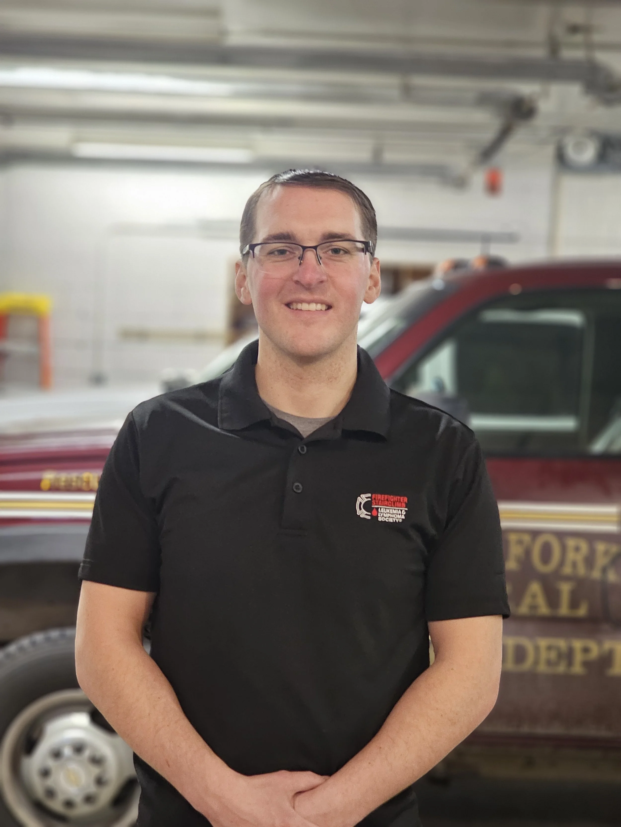 Charles Eastty, with glasses, smiling, wearing a black collared shirt with a logo in front of a fire department vehicle inside a garage.