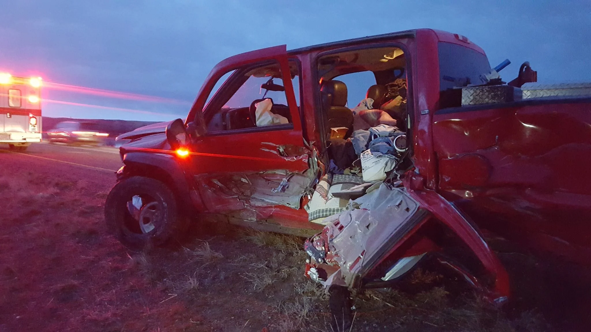 Damaged red pickup truck involved in a crash on the side of the road with emergency vehicle approaching in the background.