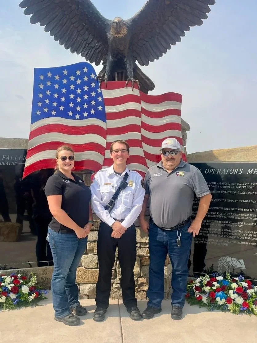 Three people standing in front of the tow memorial with a large eagle and American flags, flowers at the base, during daylight.