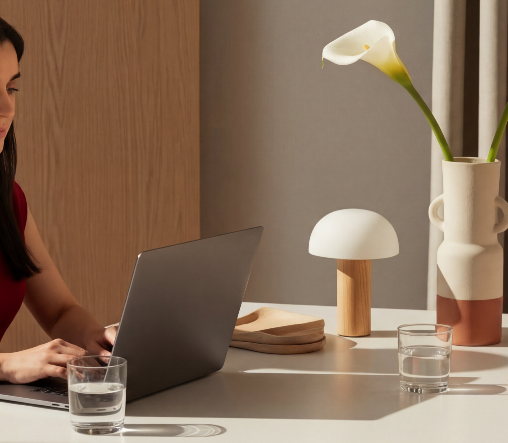 Interior scene with a woman working on a laptop at a white table, two glasses of water, a wooden tray, a mushroom-shaped lamp, and a decorative vase with a white calla lily flower.