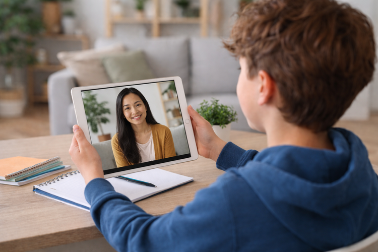 A person holding a tablet shows a woman smiling during a video call at a desk with notebooks and a pen, in a cozy living room.