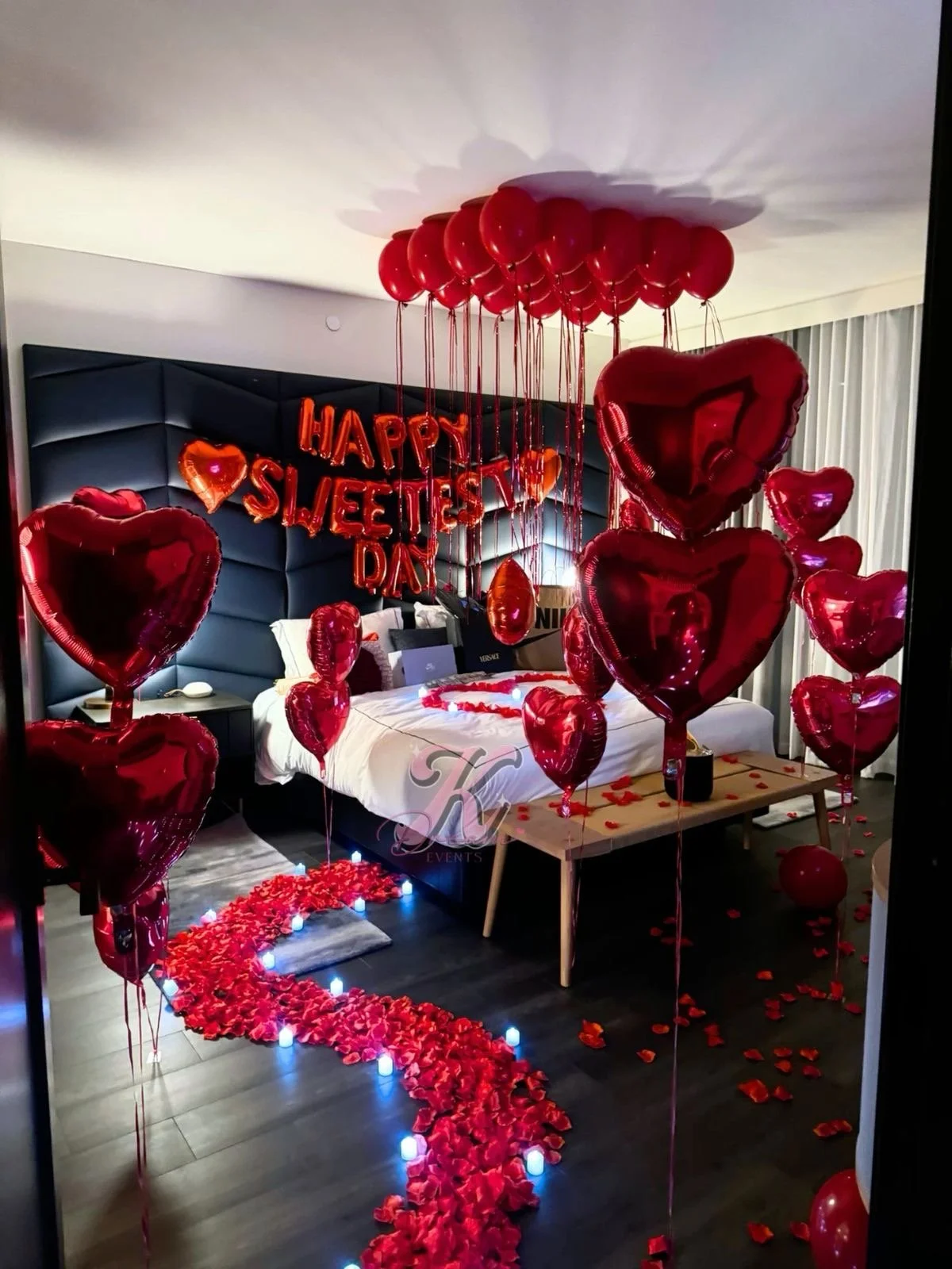 A hotel room decorated with red heart-shaped balloons, rose petals, and LED candles for a Valentine's Day celebration. A large heart-shaped balloon and a 'Happy Sweetest Day' balloon arrangement are hanging from the ceiling.