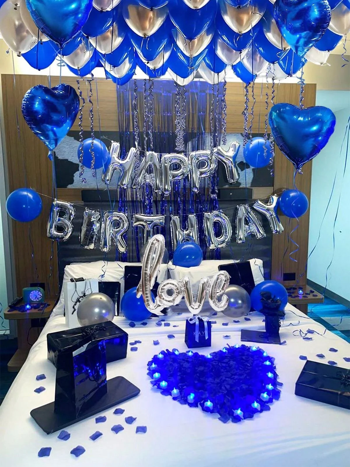 Decorated birthday celebration with numerous blue and silver balloons, silver foil letter balloons spelling 'HAPPY BIRTHDAY' and 'love', a blue heart-shaped light-up decoration, gift boxes, confetti, and LED heart-shaped lights on a table.