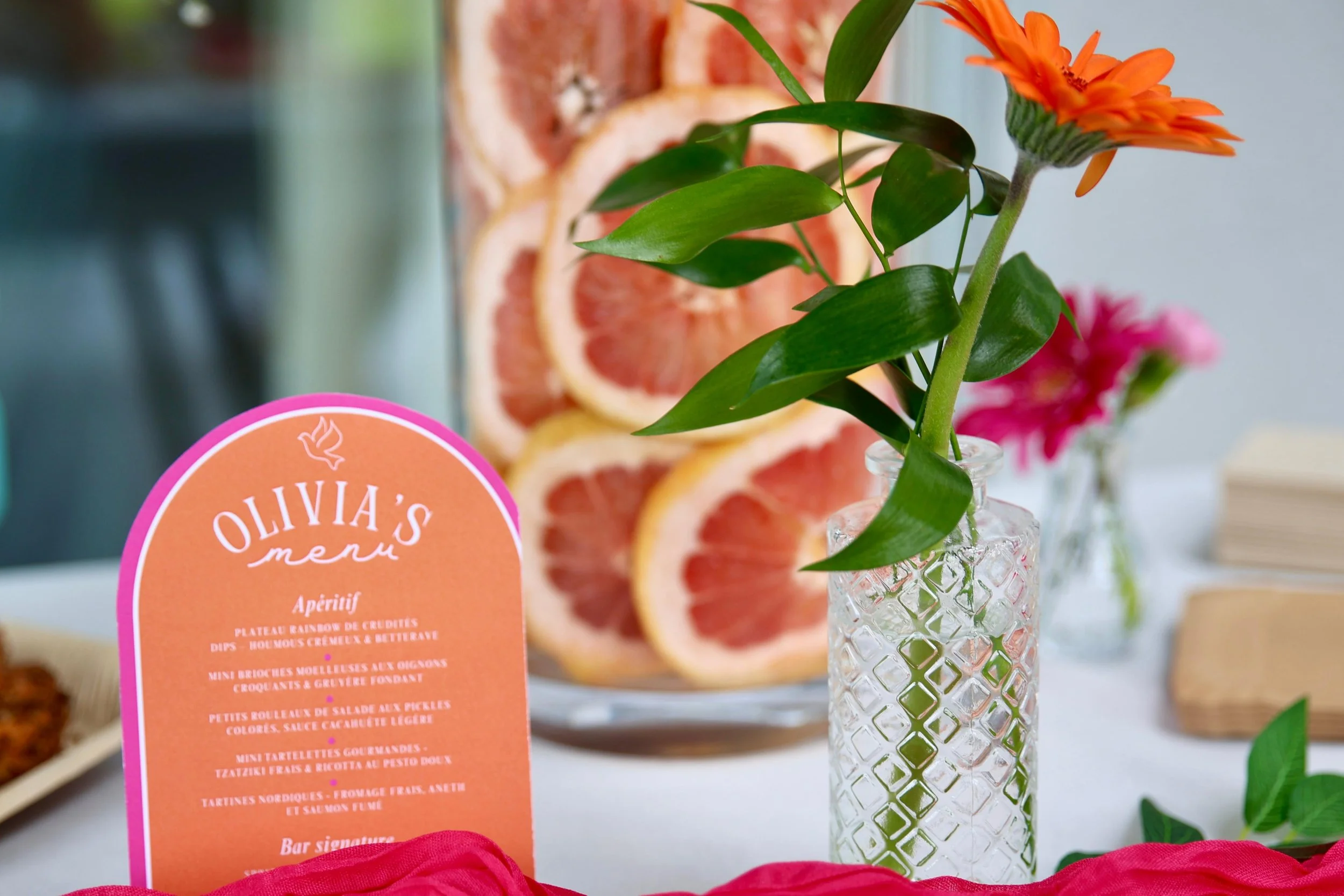 Pink menu card in front of a vase with green leaves and orange flowers, with slices of grapefruit and other food items visible in the background.