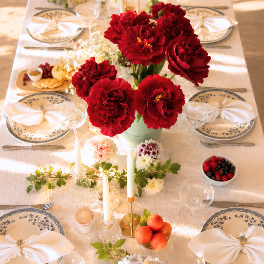 A beautifully decorated dining table with a large bouquet of red flowers as the centerpiece, surrounded by plates with white napkins, glasses, and various bowls of berries and peaches, set for a meal.