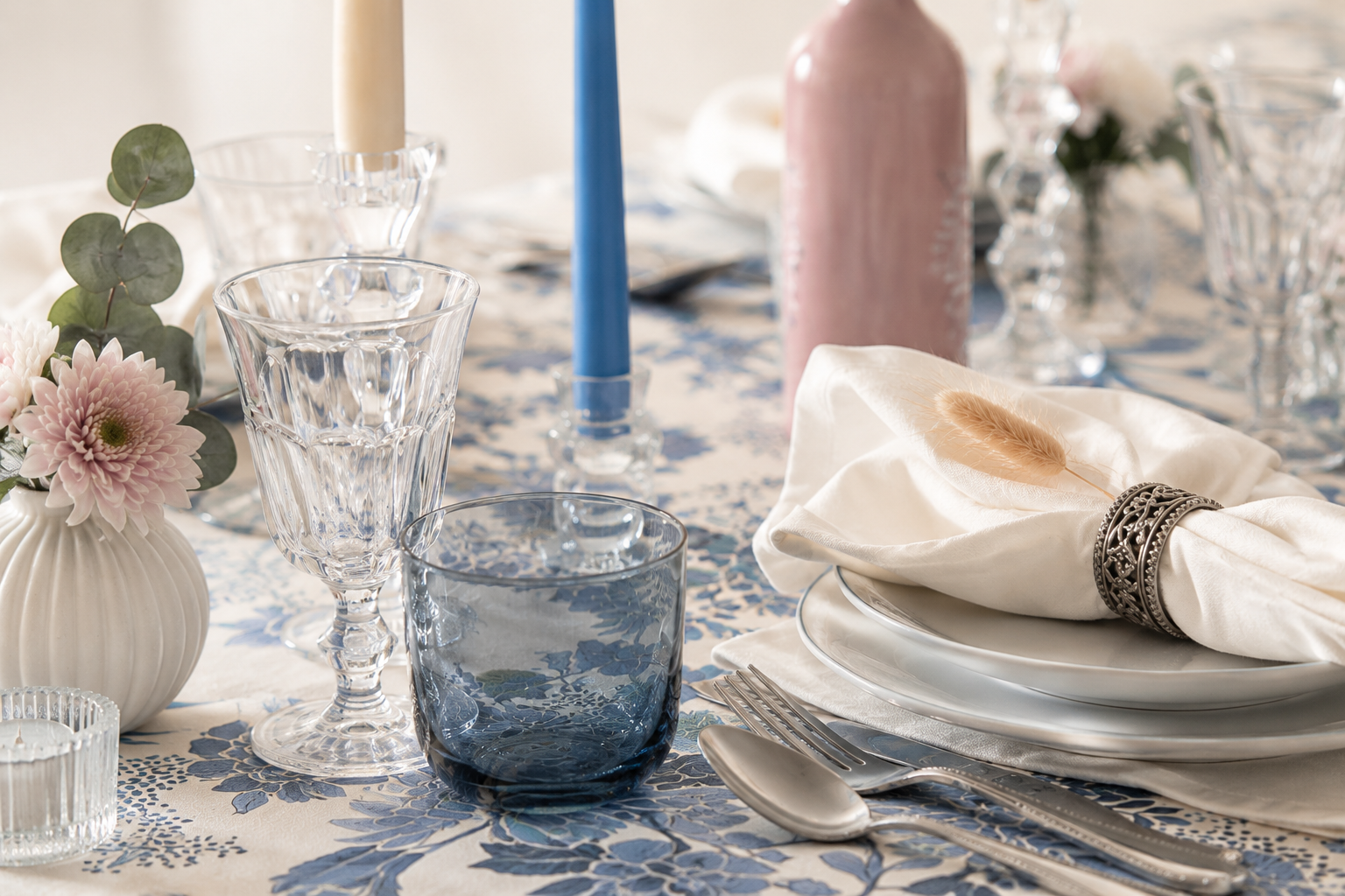 Elegant table setting with glassware, floral arrangements, candles, and folded napkins on a blue patterned tablecloth.