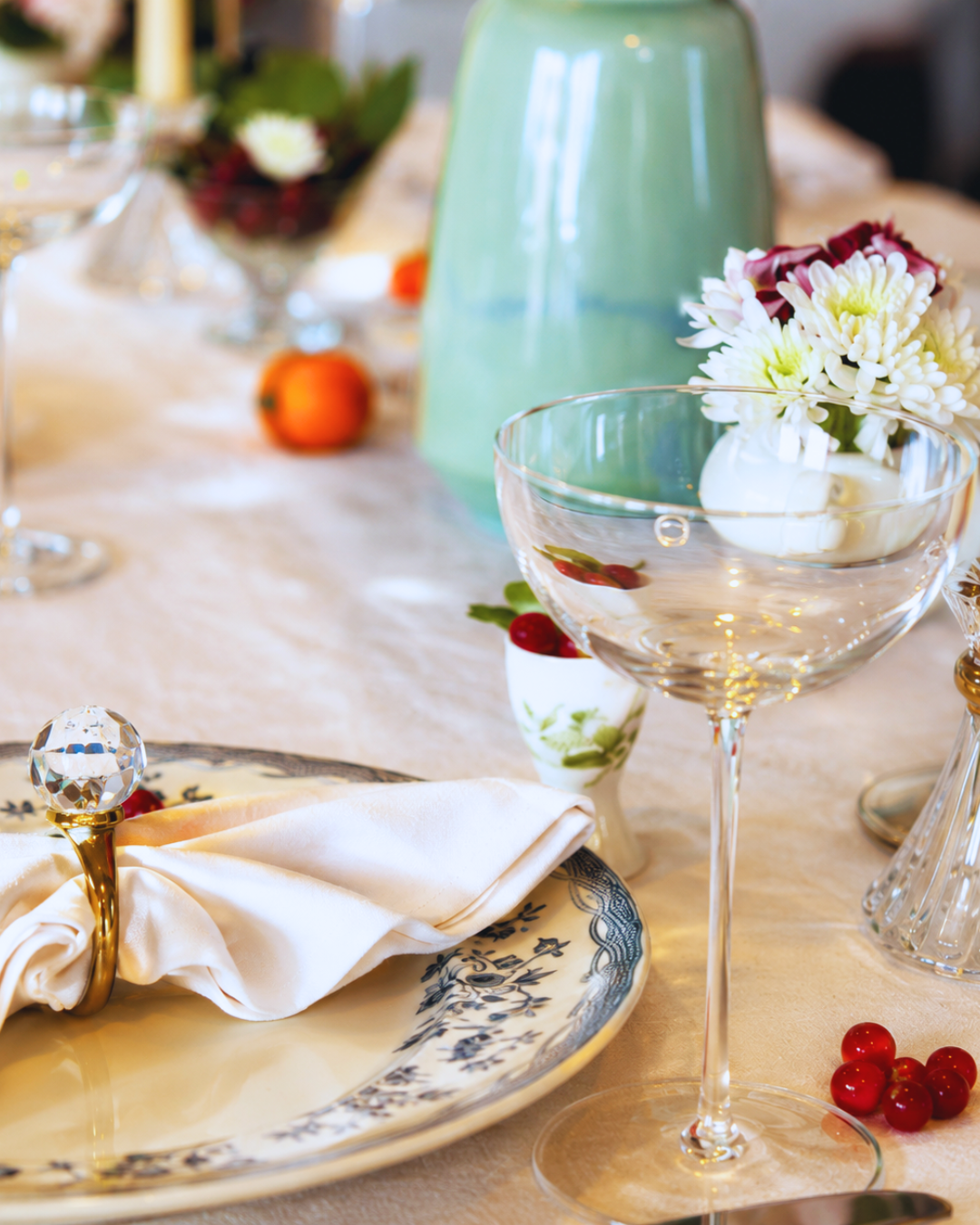 Elegant table setting with vintage patterned plate, crystal glassware, white napkin with decorative gold napkin ring, and floral centerpiece with white and dark pink flowers in a white vase.