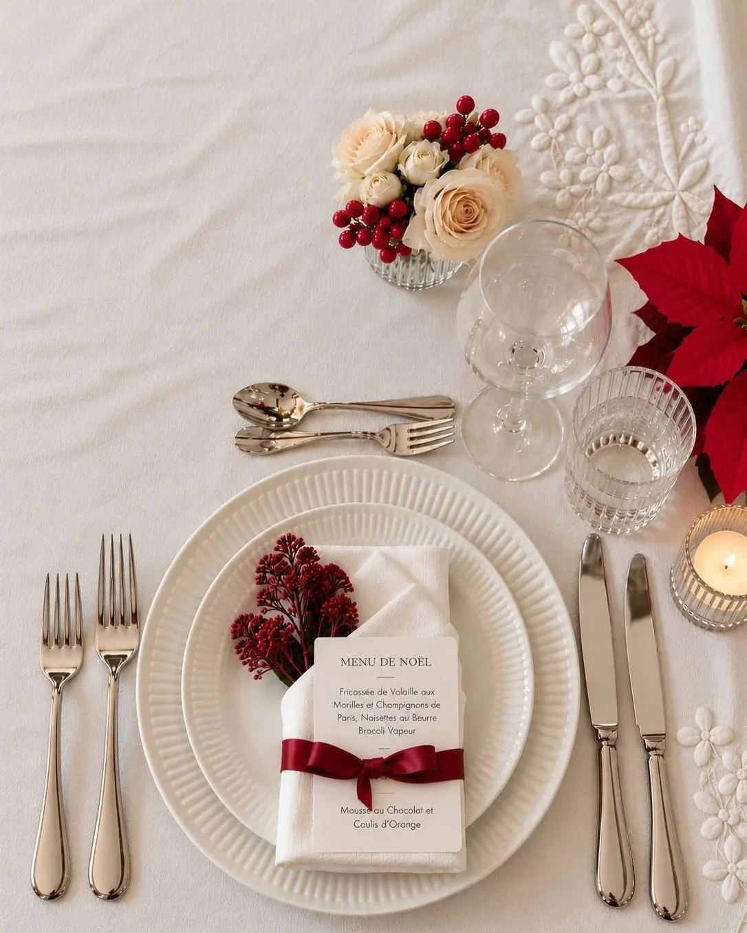 Elegant holiday table setting with white plates, silverware, wine glass, water glass, candle, poinsettia, and a decorated napkin with a menu card, red ribbon, and red flower.