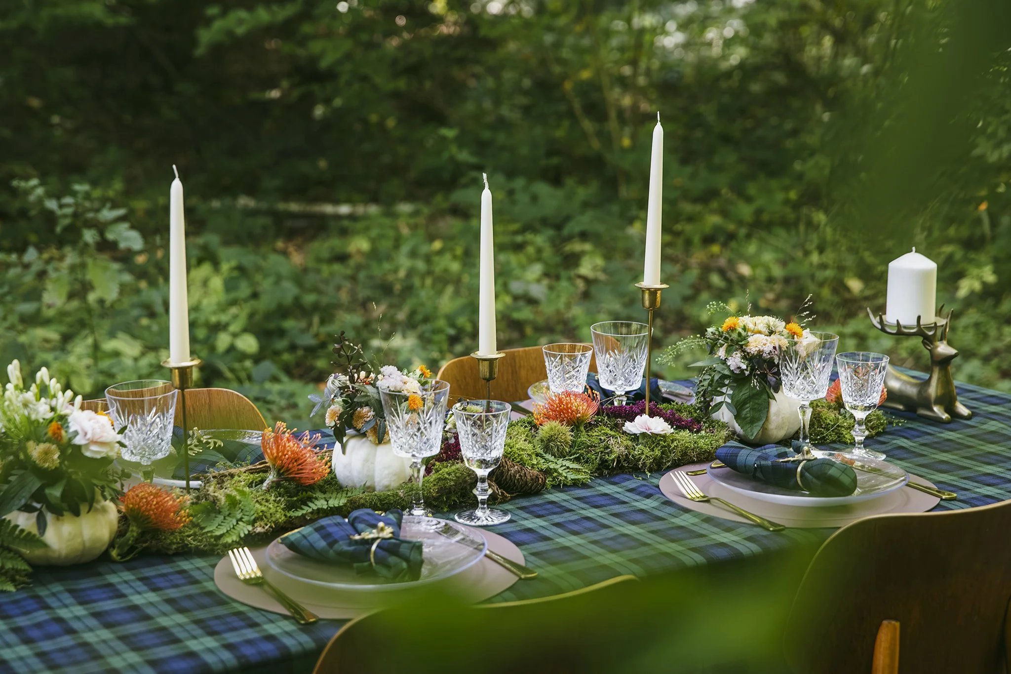 Outdoor table setting with candles, flowers, and glassware on a green tartan tablecloth, surrounded by greenery.