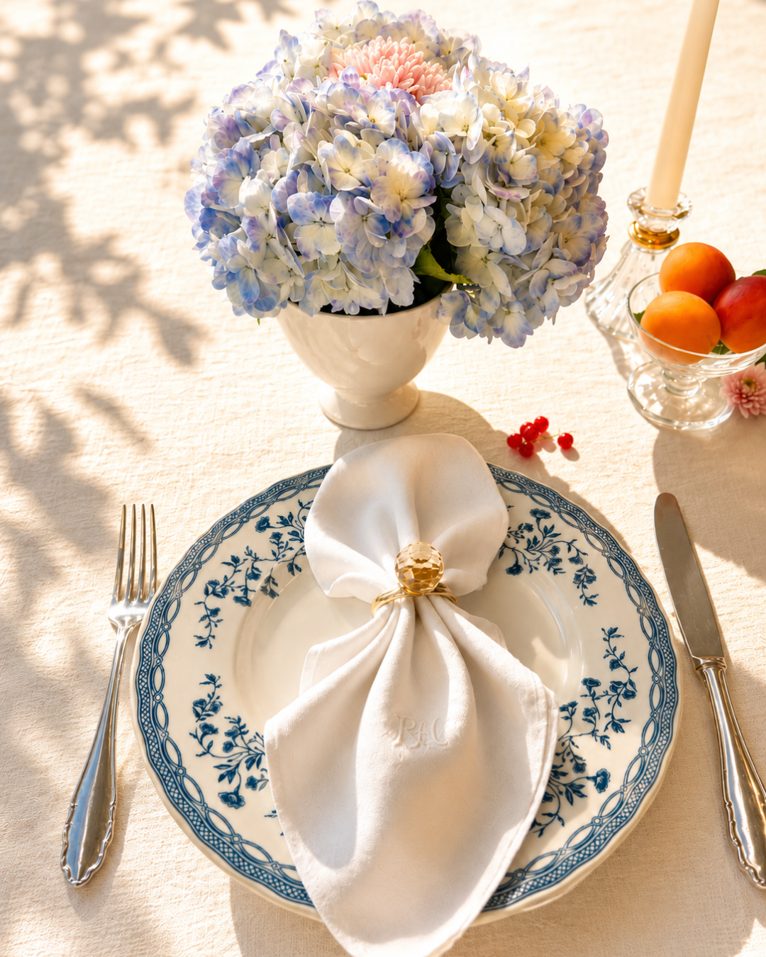 A formal dining table setting with a blue and white floral plate, silverware, a white napkin secured with a gold ring, a blue and white floral centerpiece with hydrangeas, a glass bowl with peaches, and a white candle in a glass holder, all on a cream tablecloth with natural sunlight.