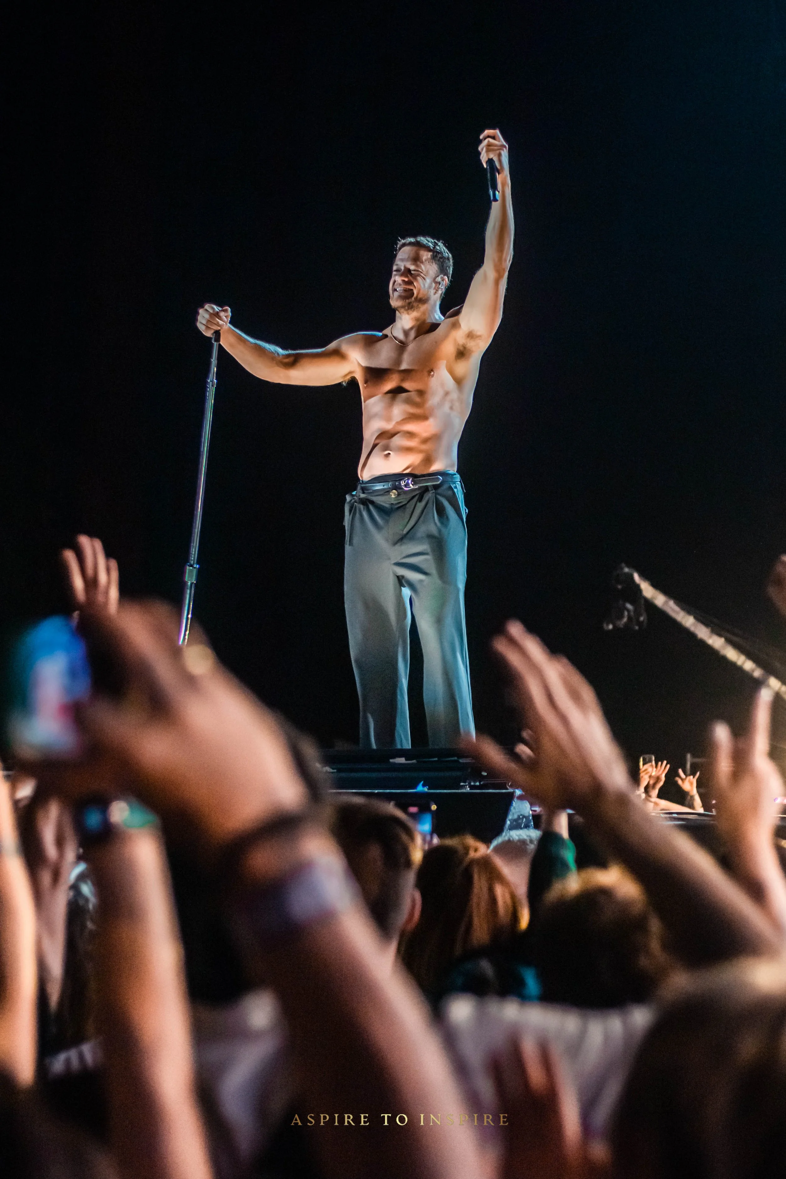 A shirtless male performer on stage with no shirt, wearing dark pants, holding a microphone in one hand and raising the other hand. The performer is smiling and the audience's hands are raised toward him in the foreground.