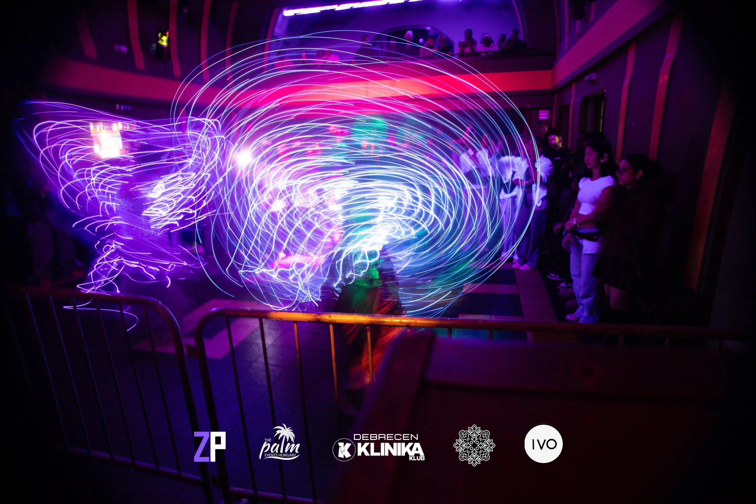 People watching a light performance with swirling purple, pink, blue, and green lights in a dark indoor venue, with logos at the bottom for ZP, The Palm Events Hungary, Debrecen Klinika, an intricate snowflake design, and IVO.