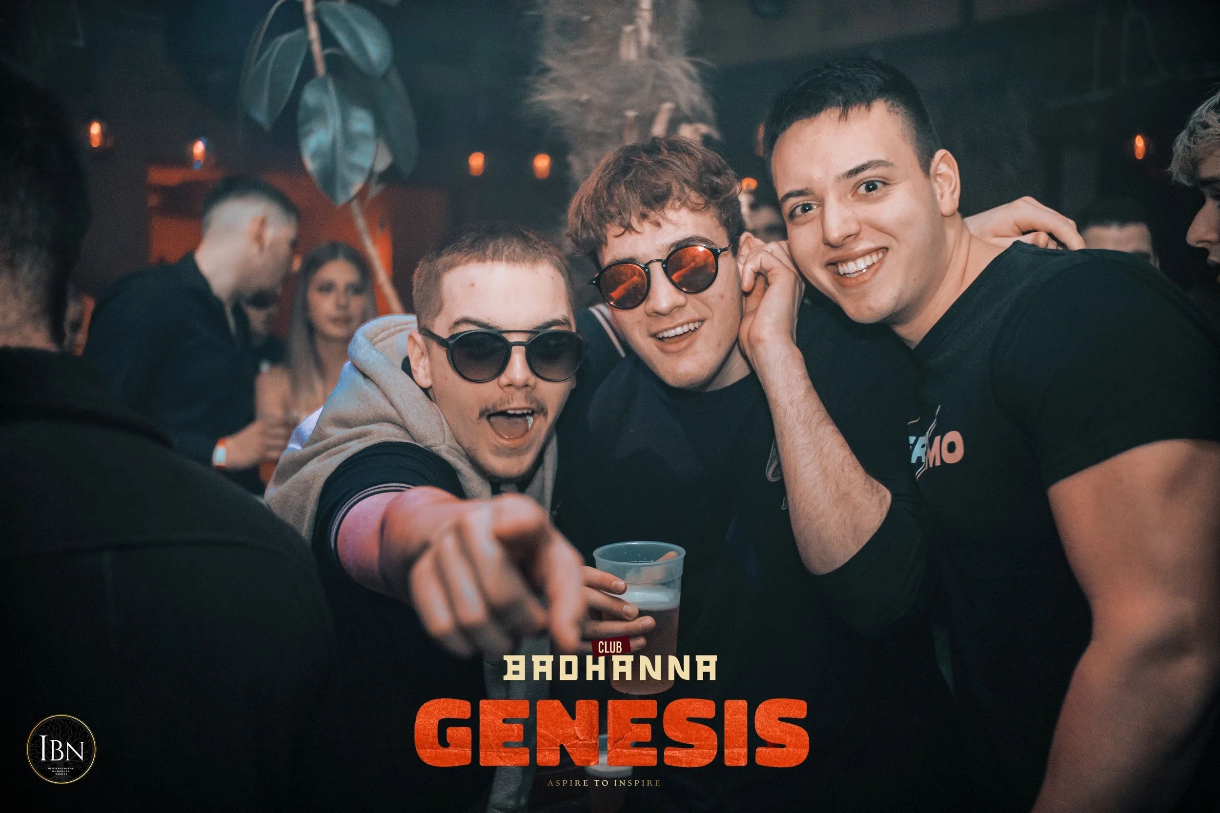 Three young men wearing sunglasses smiling and partying at a nightclub with a crowd in the background, and a logo for 'Badhanna Genesis' at the bottom.
