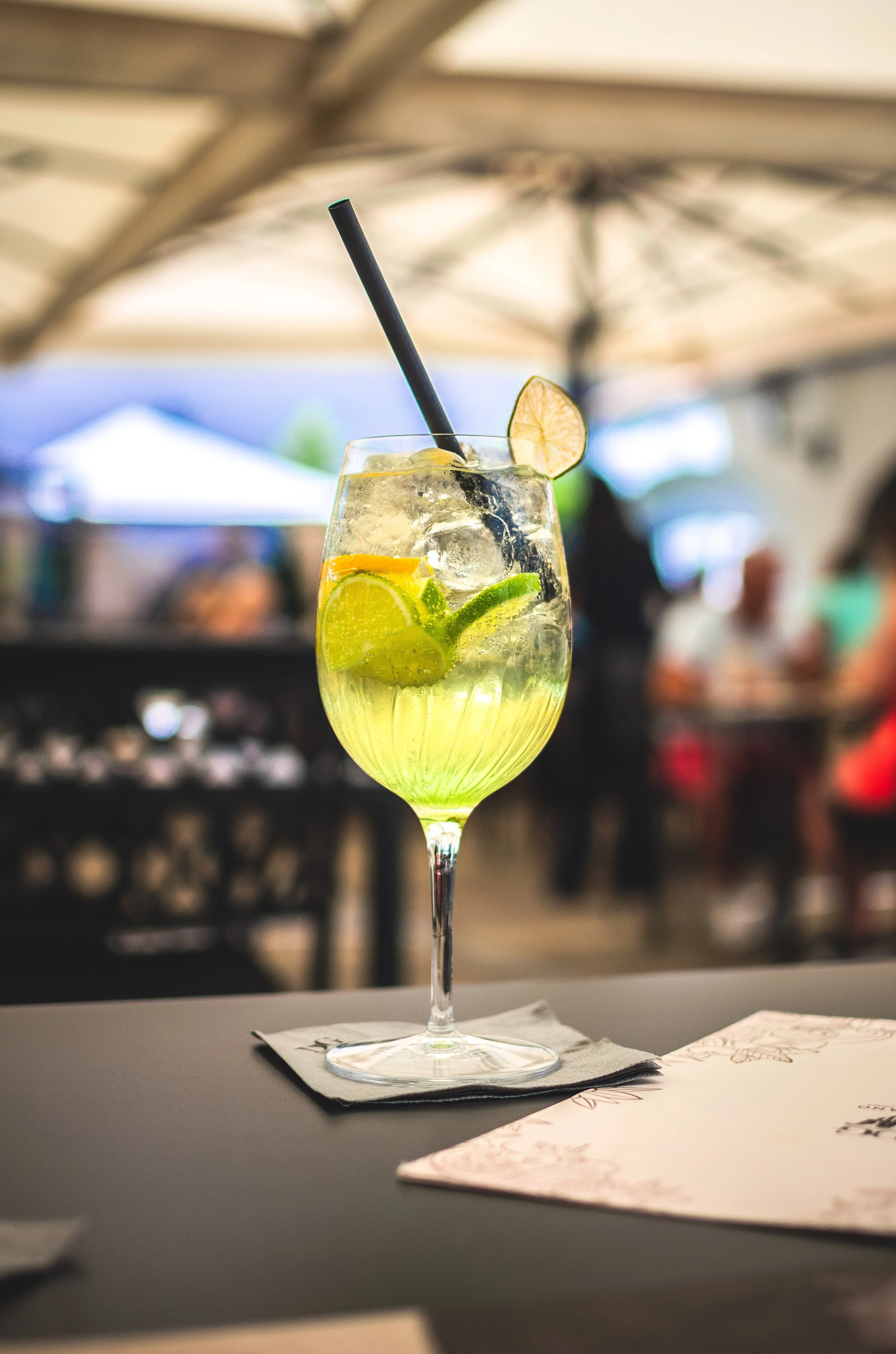 A cocktail glass with lime, lemon, and lime wedges, ice, and a straw, garnished with a lemon slice, set on a napkin at an outdoor dining area.