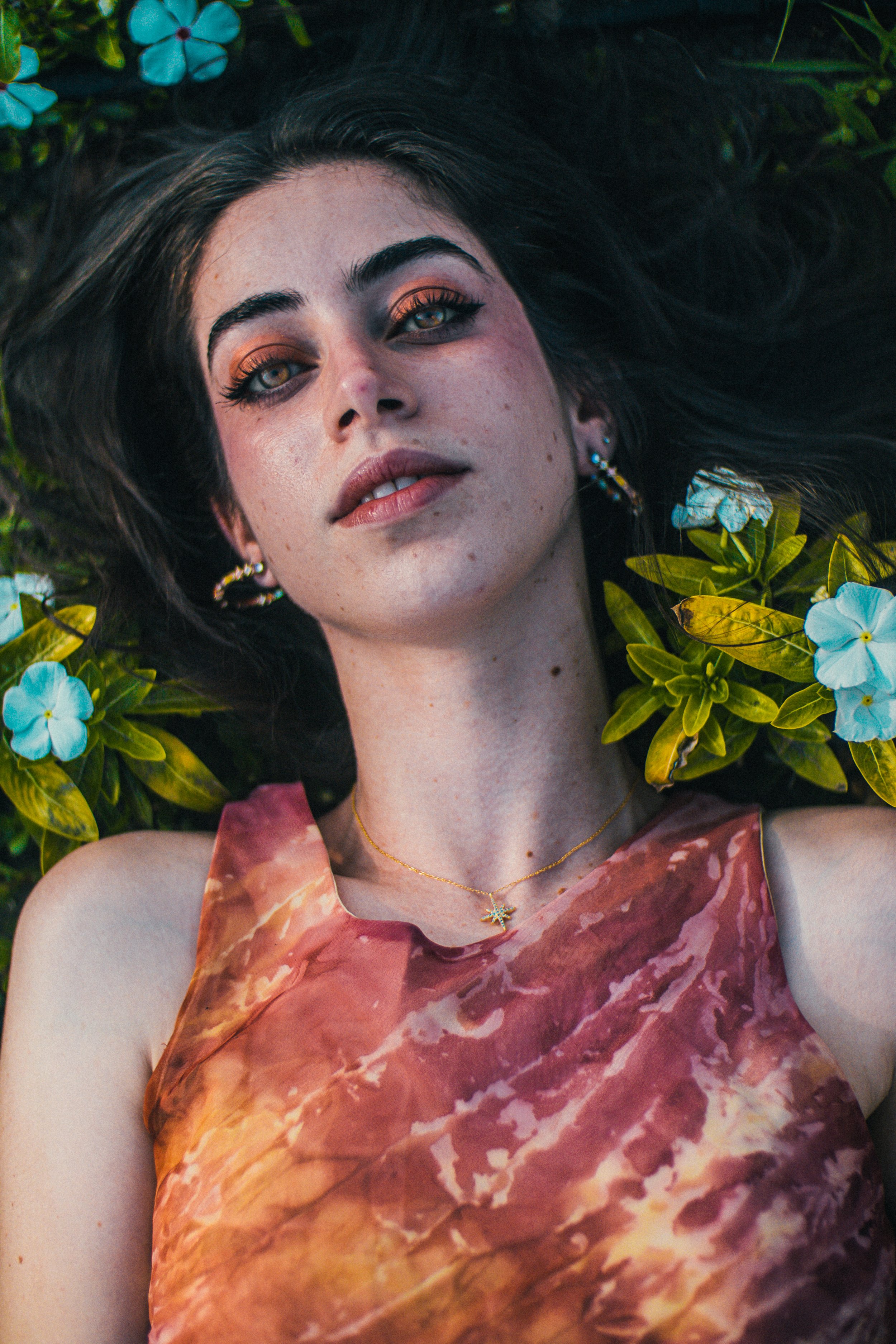 A young woman with brown hair and brown eyes lying on the ground, surrounded by green leaves and light blue flowers, wearing a pink and orange tie-dye sleeveless top and a small star-shaped necklace.