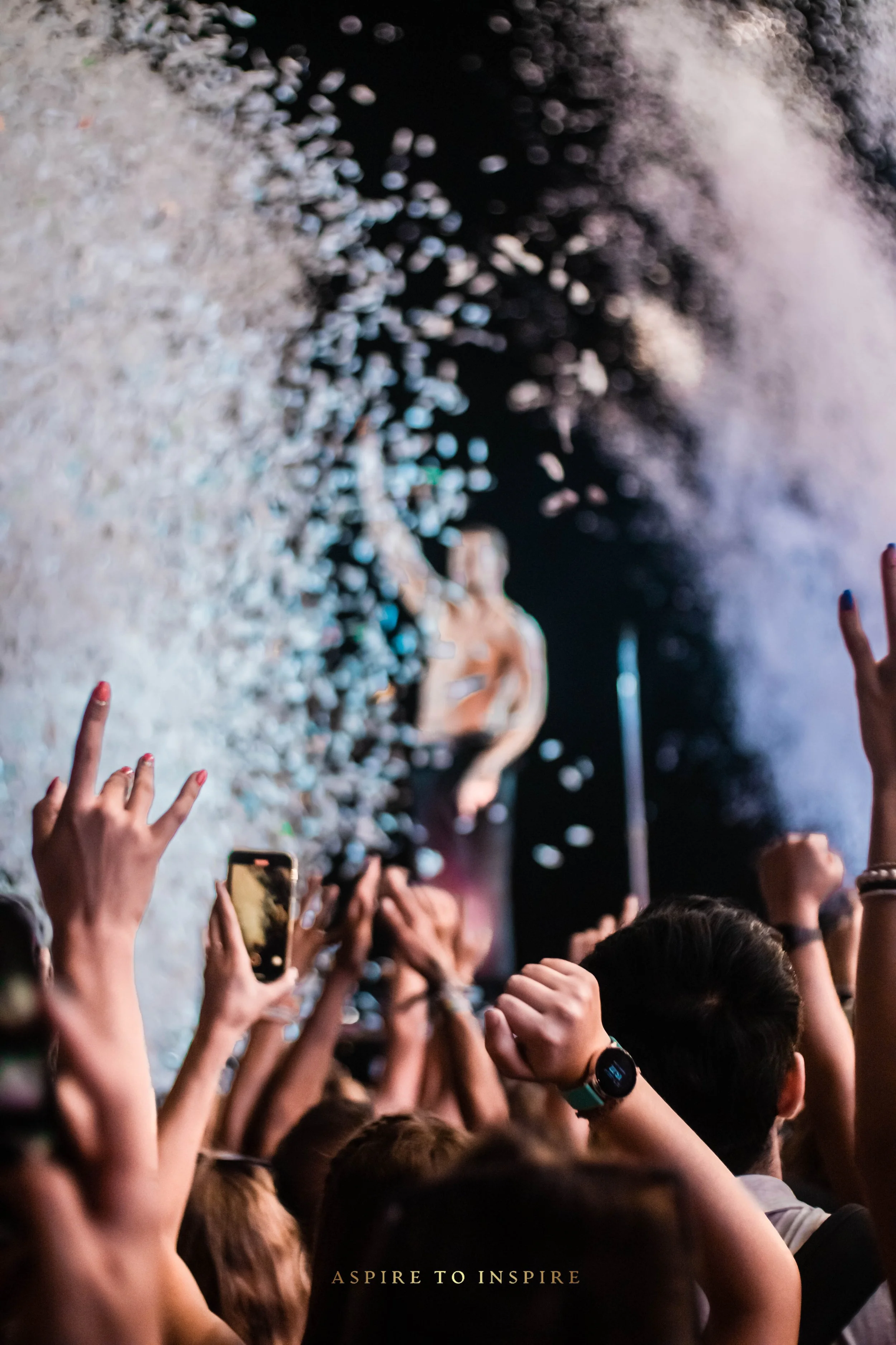 Concert crowd with hands in the air, confetti falling, and a singer on stage with fog and lights, with the phrase 'Aspire to Inspire' at the bottom.