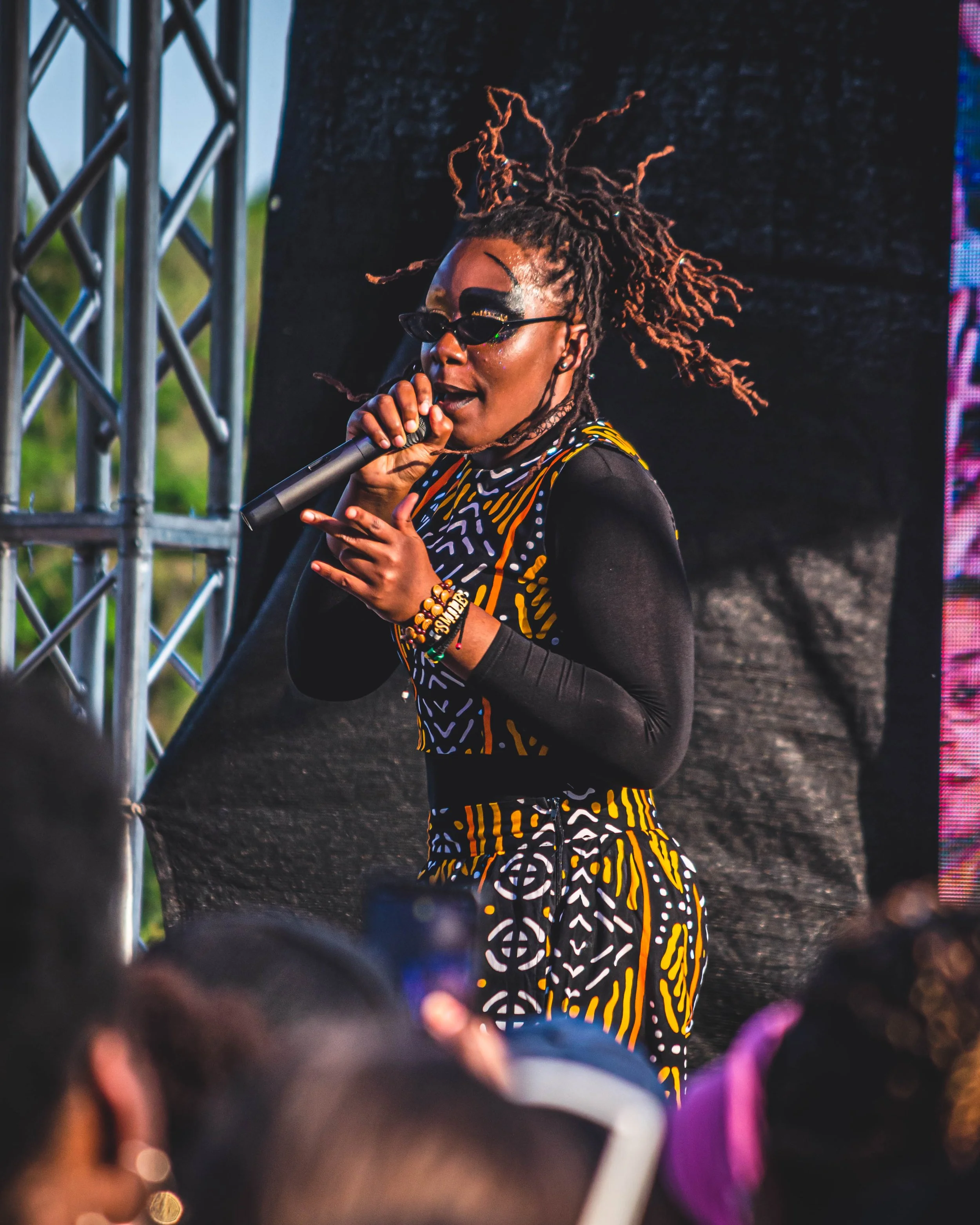 A woman with dreadlocks, wearing sunglasses and a colorful patterned outfit, singing into a microphone on stage at an outdoor concert.