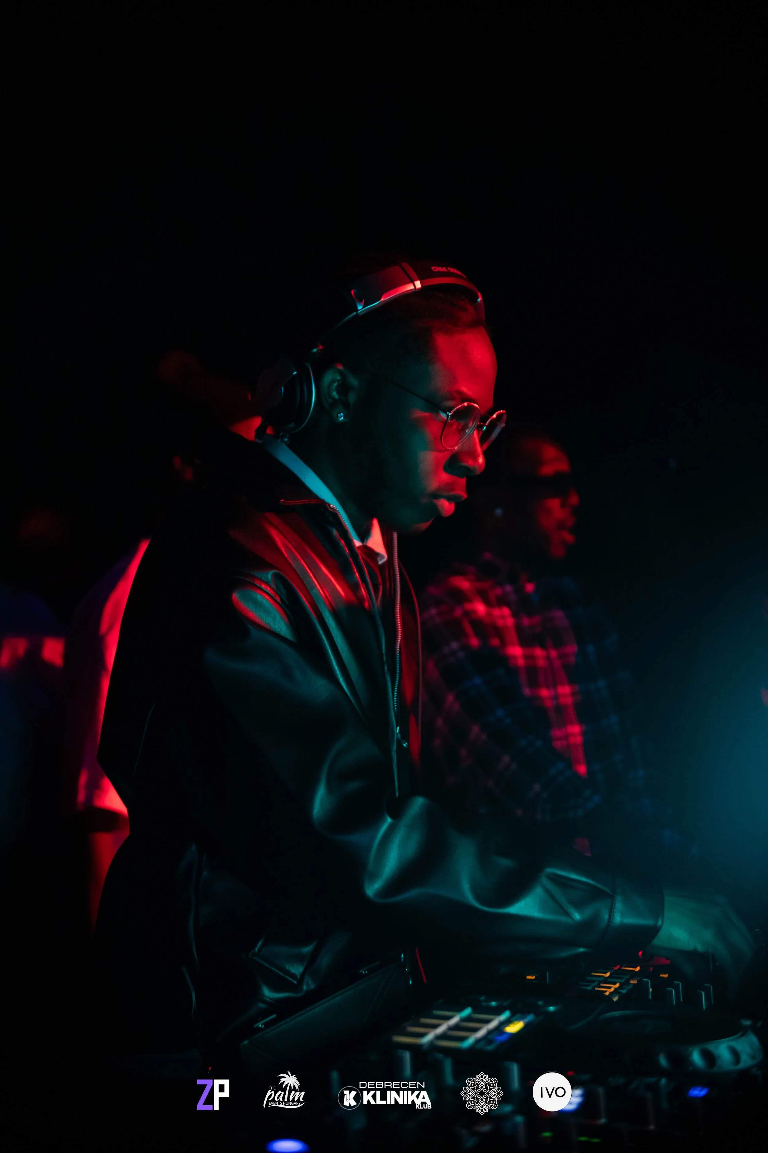A DJ wearing headphones, glasses, and a leather jacket, performing at a nightclub with a dark background and neon lighting.