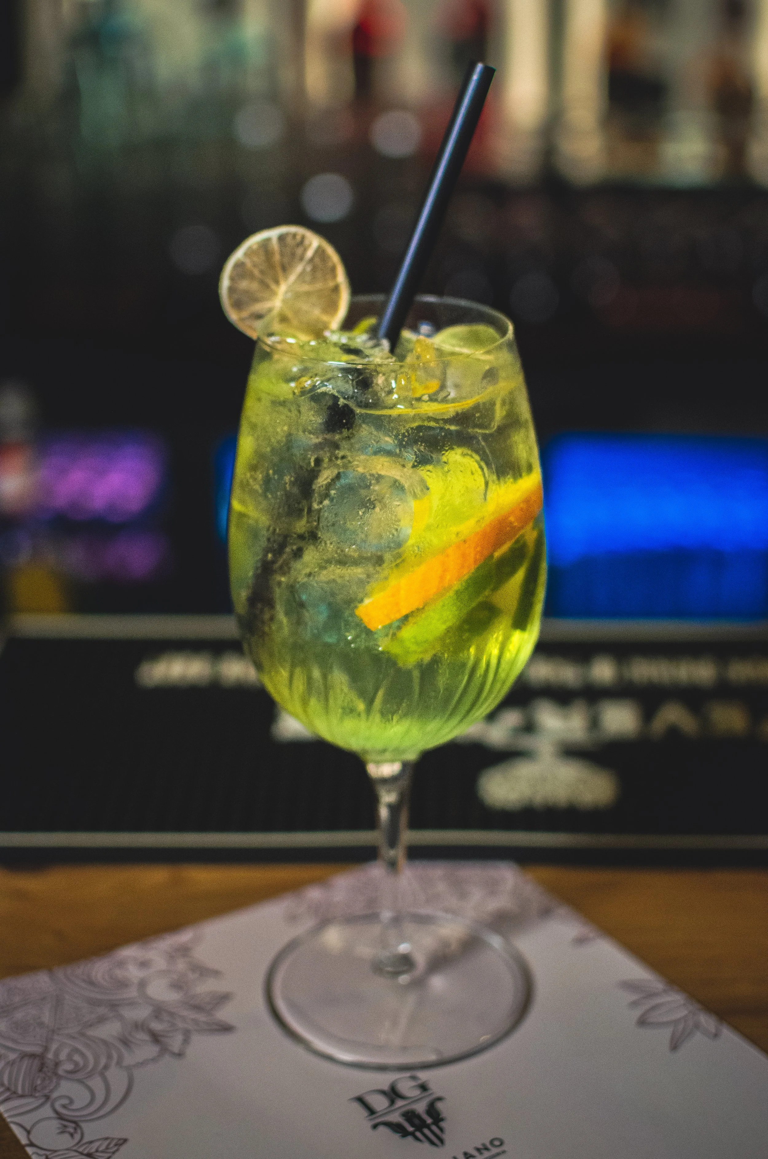 A glass of lime and cucumber cocktail with lemon slices, a black straw, and ice cubes on a bar counter.