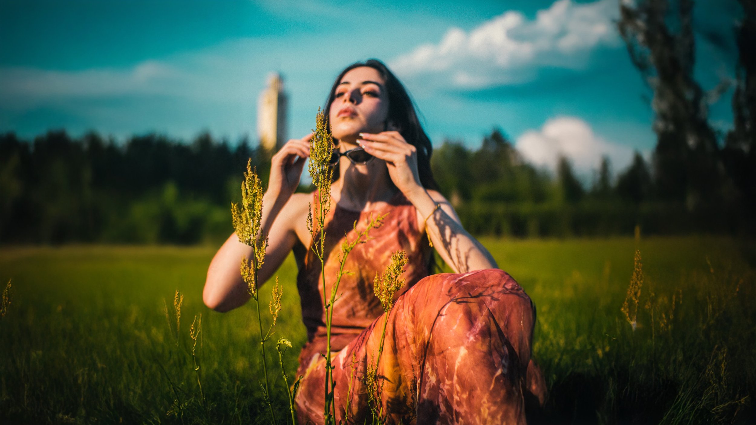 A woman with dark hair wearing an orange and pink patterned dress sits on the grass in a green field, holding sunglasses near her face, with tall yellow wildflowers in front of her. The background shows a blue sky with clouds and a distant tower.