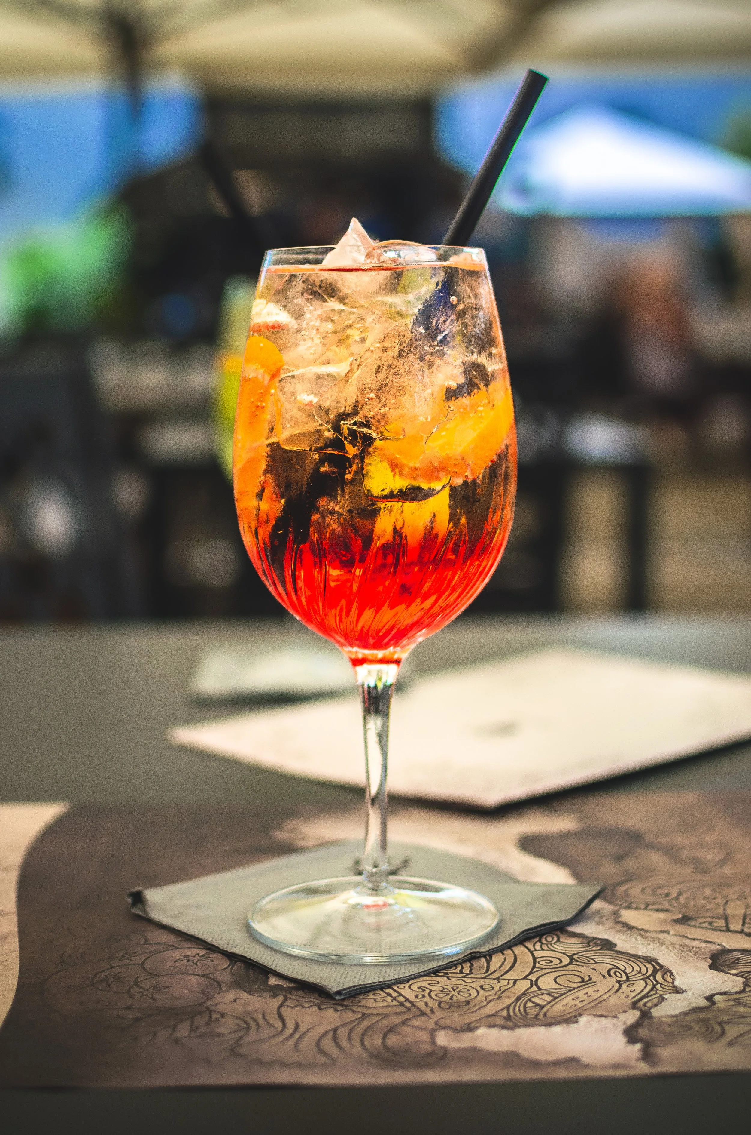 A tall glass with a red and orange layered cocktail, ice, and orange slices, garnished with a black straw, on a napkin on a table outdoors with blurred umbrellas and greenery in the background.