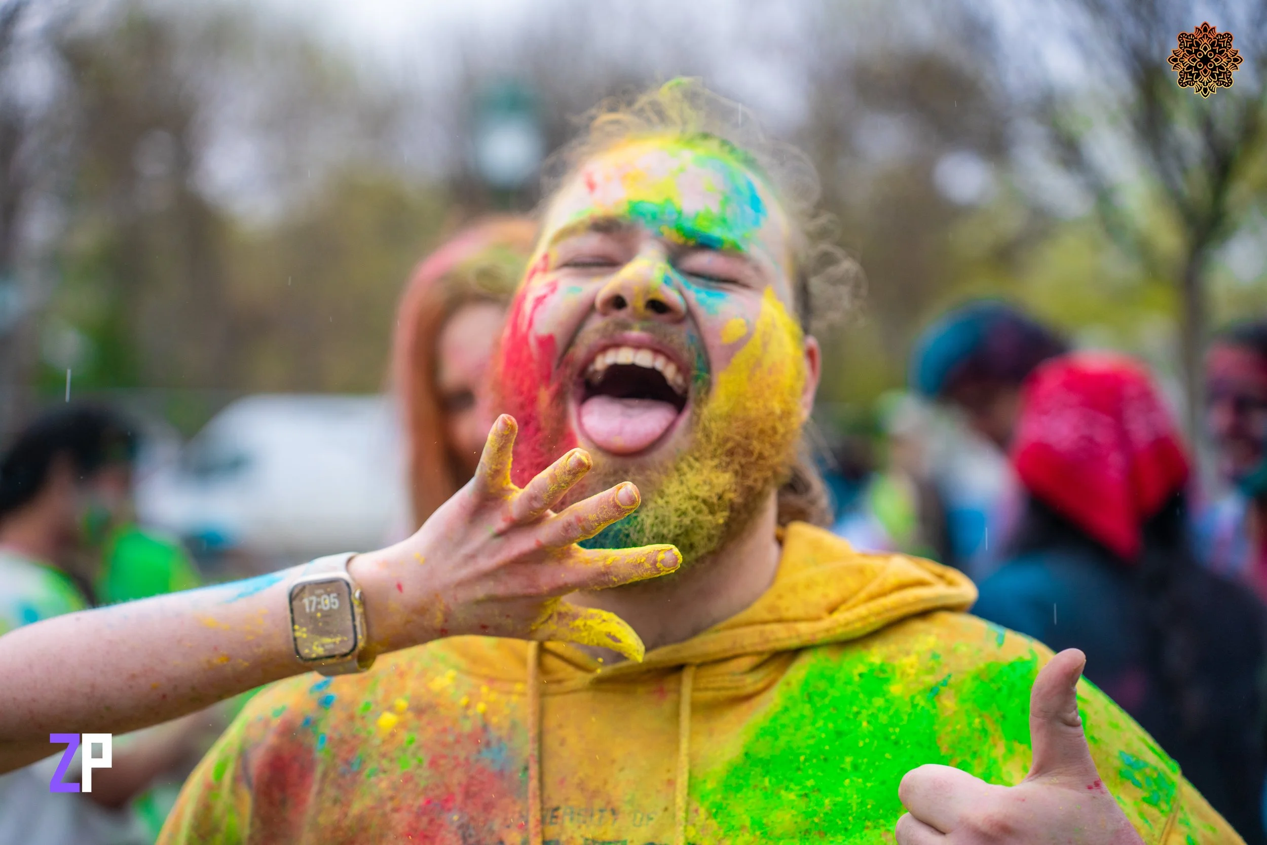 Person covered in yellow, green, red, and blue powders celebrating at a color festival outdoors, smiling and giving a thumbs-up.