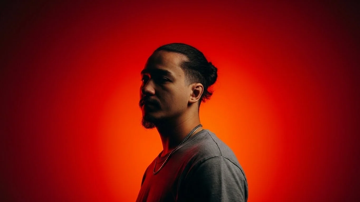 A man with a mustache and hairstyle tied back, wearing a gray t-shirt and chain, stands in front of a red-orange gradient background, looking to the side.
