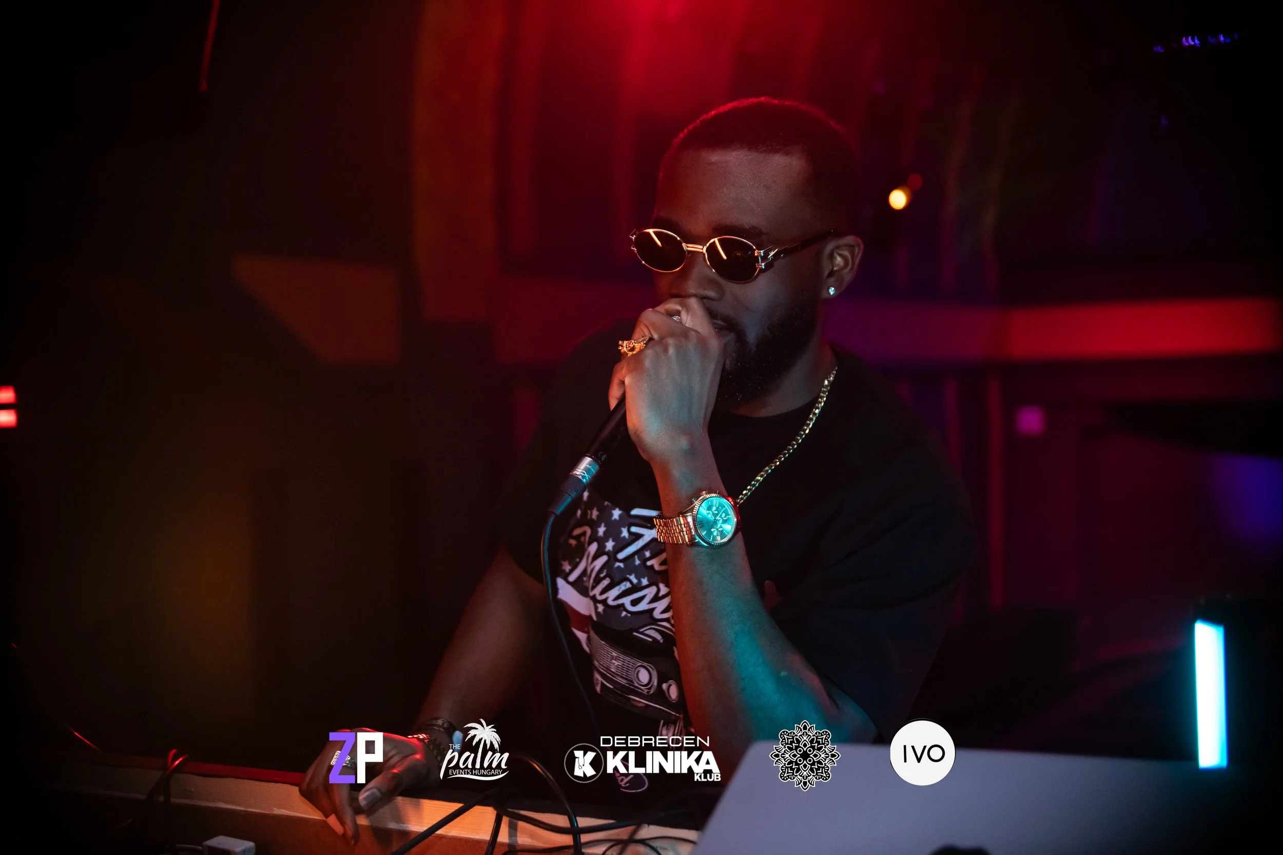 A DJ with sunglasses, gold watch, gold chain, and earrings DJing in a dark club with red and blue lighting.