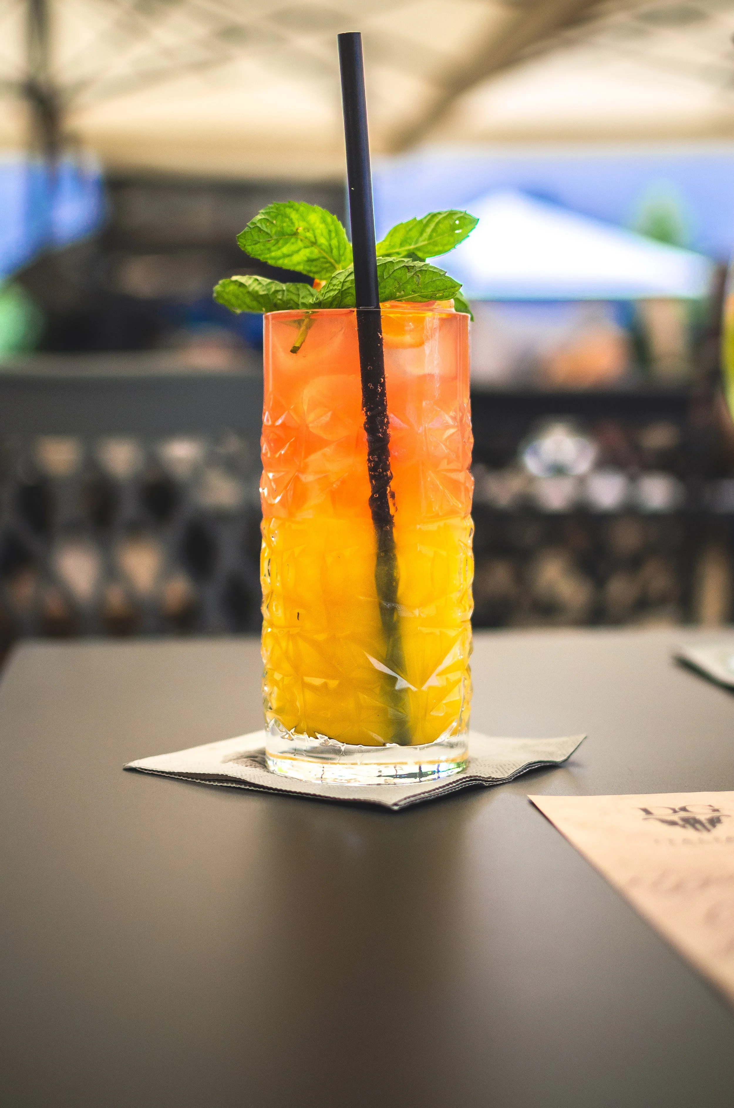 A layered colorful cocktail in a tall, textured glass garnished with fresh mint leaves and a black straw, placed on a napkin on a dark table.