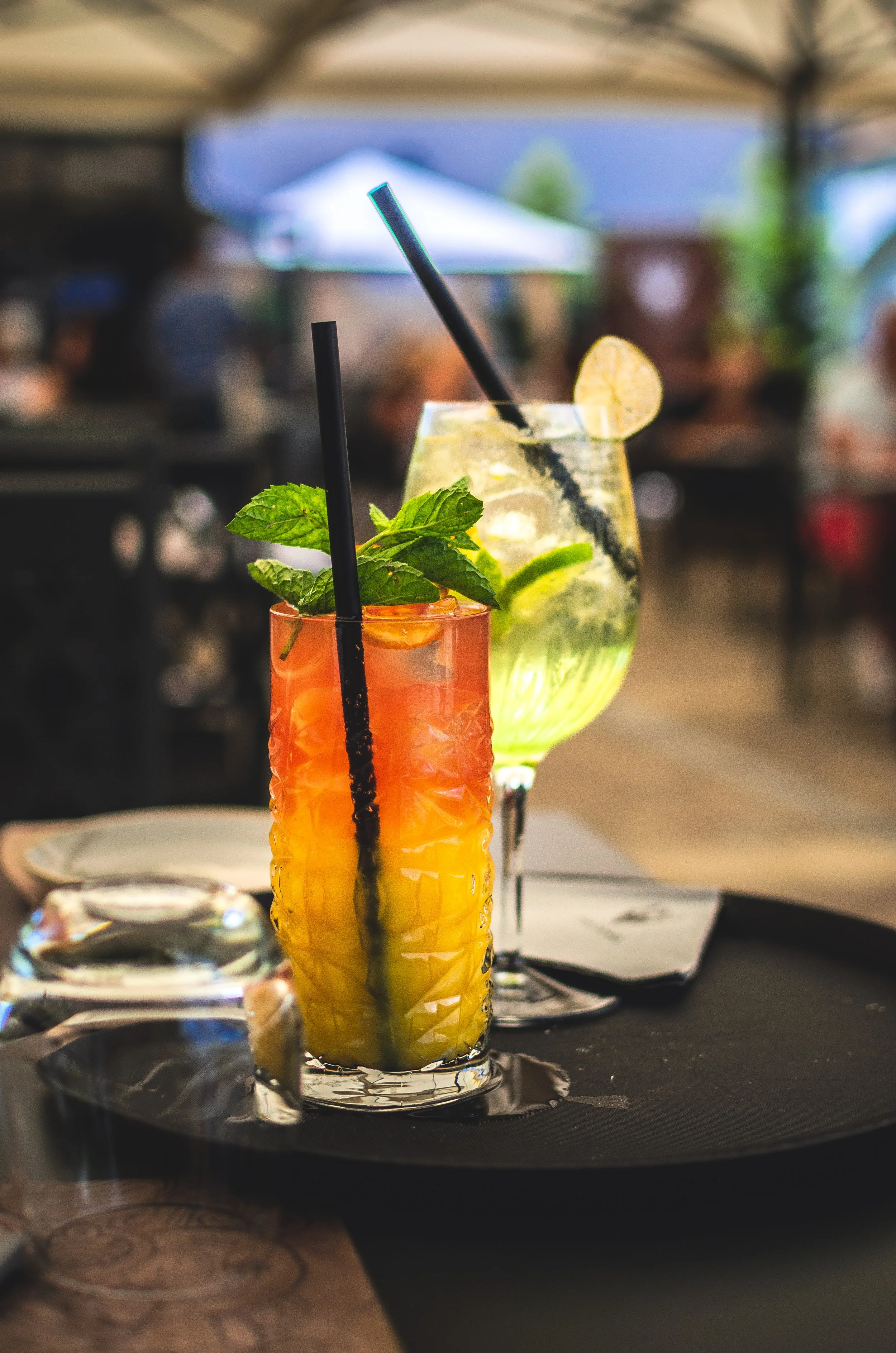 Two colorful cocktails on a black table, one with layered yellow and orange colors garnished with mint and a black straw, the other with lime slices and ice, garnished with a lemon wedge and a black straw, in an outdoor setting.