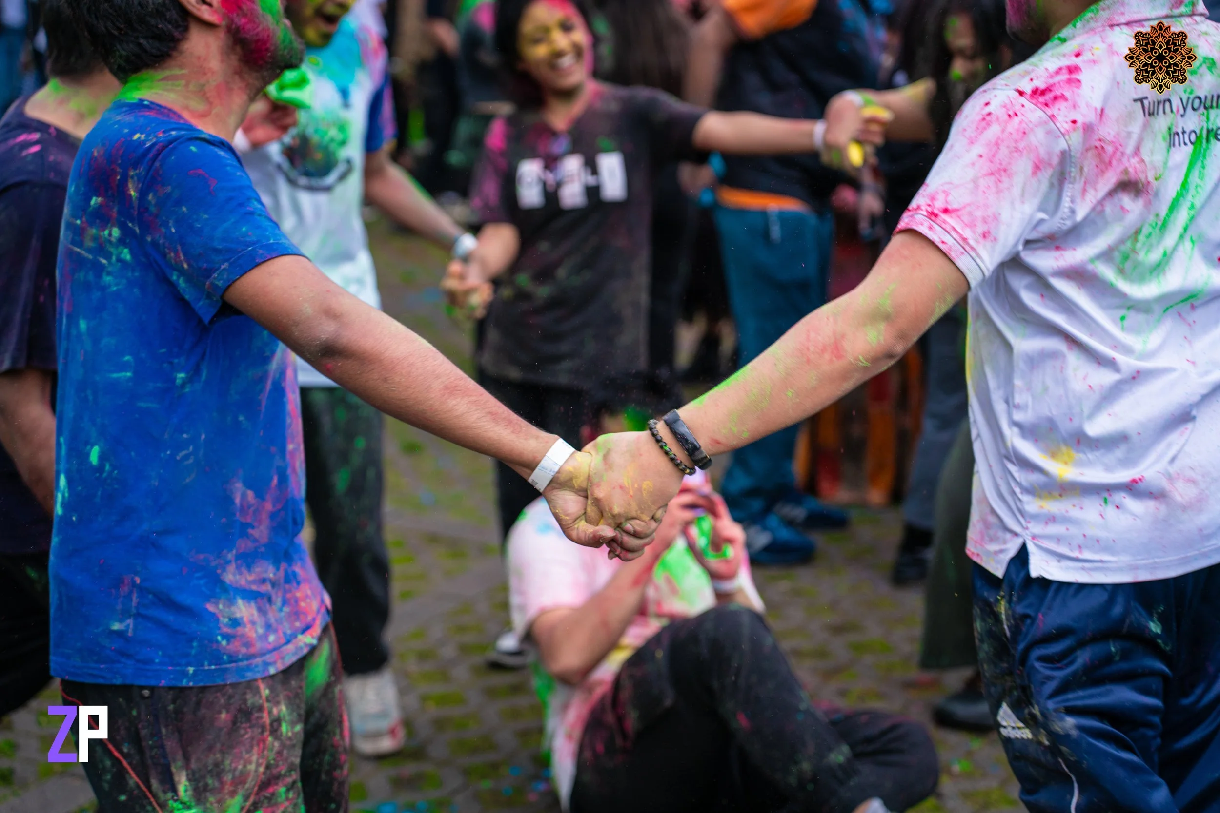 Two young people holding hands at a color run event, covered in colored powder, with others smiling and celebrating in the background.