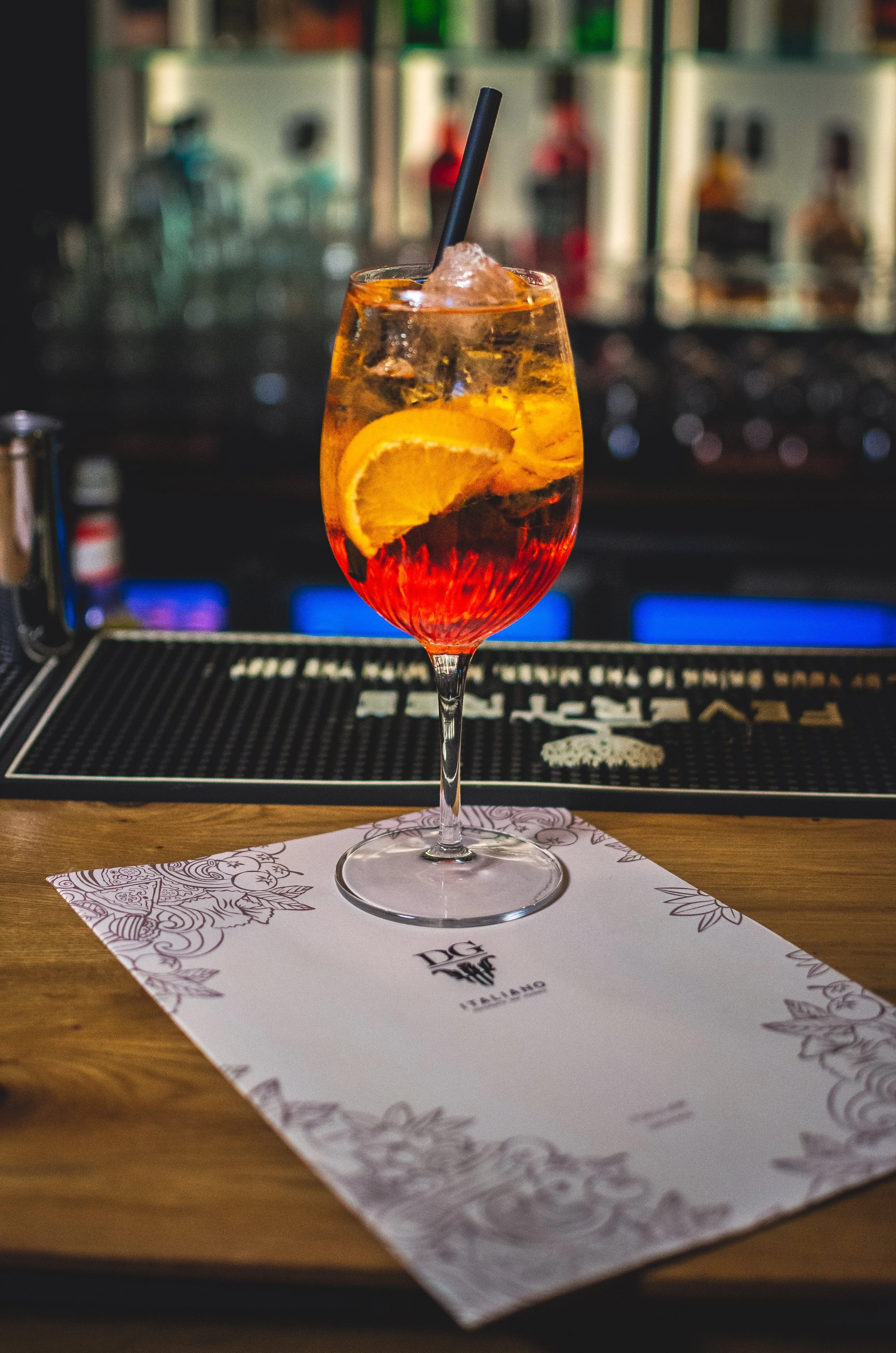 A cocktail glass filled with an orange and red drink, garnished with an orange wedge and ice cubes, sitting on a bar counter with a black straw. The background is blurred, showing shelves with bottles.