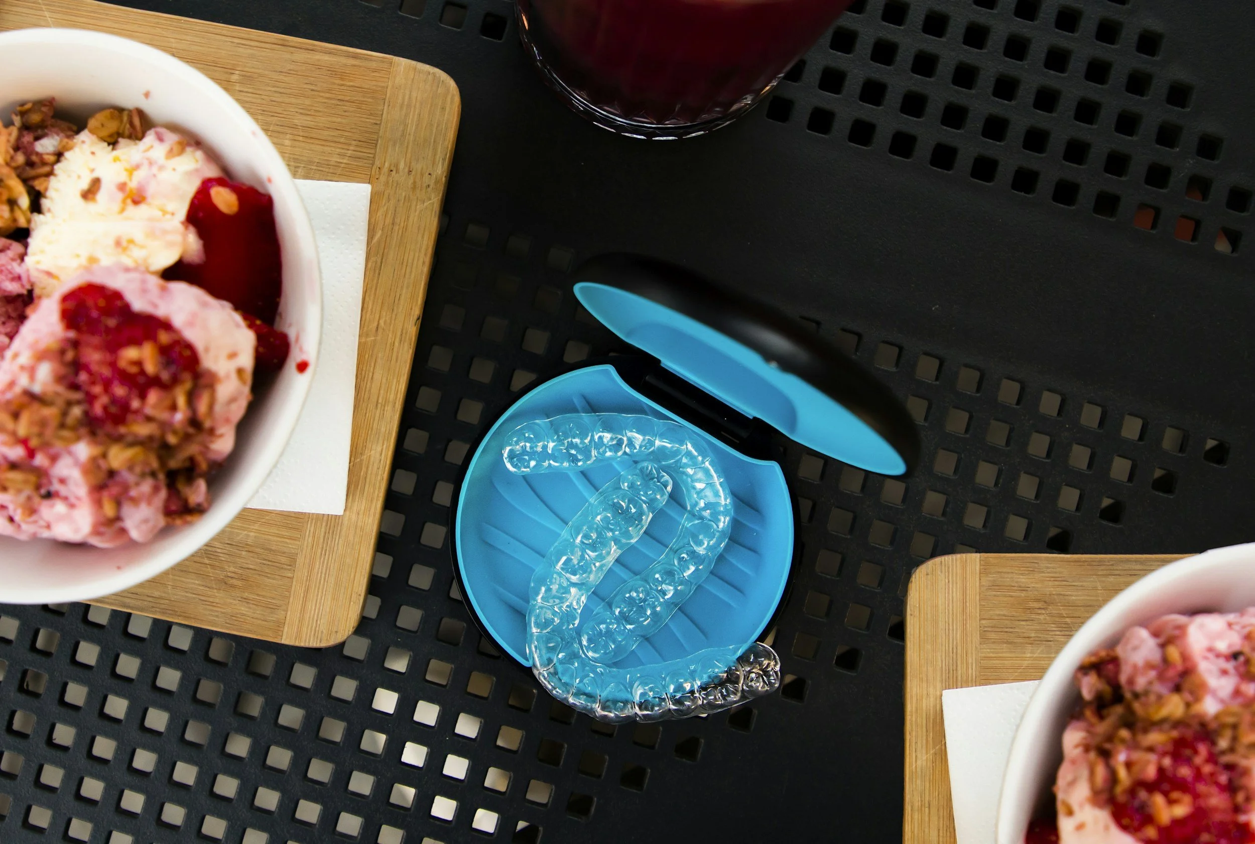 Caja de alineadores invisibles Invisalign con placas transparentes colocadas en su interior sobre una mesa de plástico negro y dos platos con helado y frutas. Los alineadores pueden removerse para comer cualquier comida.