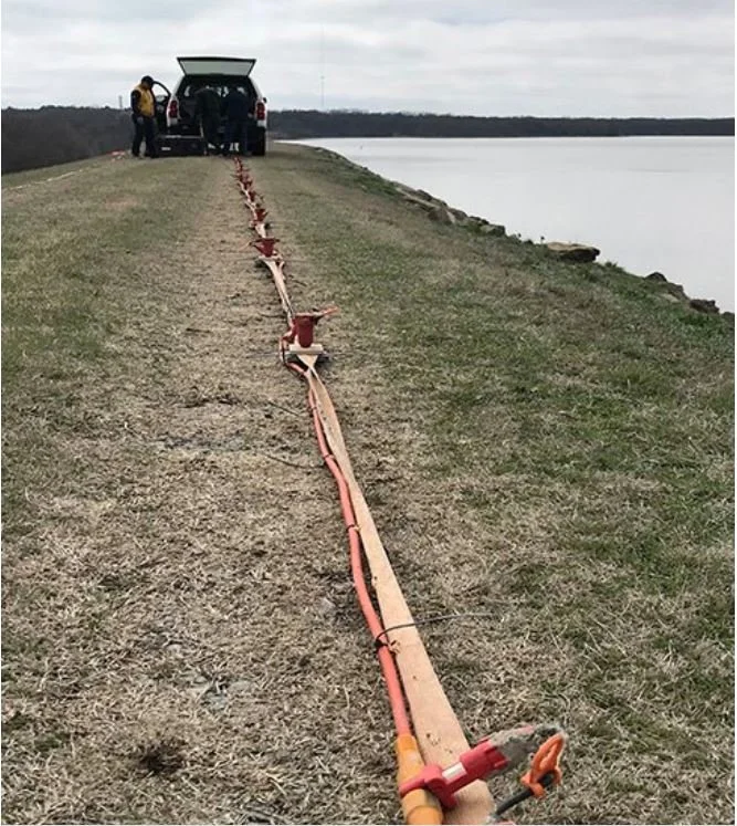 A long orange pipe with equipment attached is laid out on the ground near a water body, with a group of people and a vehicle at the far end.