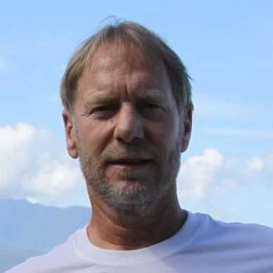 A middle-aged man with light brown hair and a beard outdoors against a blue sky with clouds.