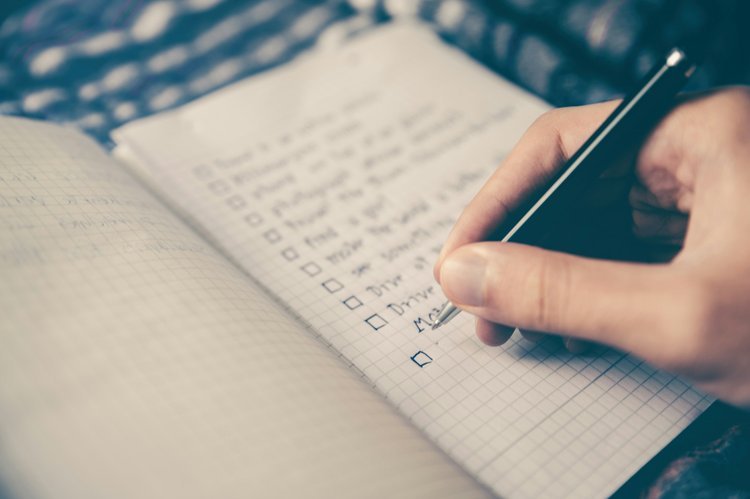 A person writing a checklist in a grid notebook with a black pen.