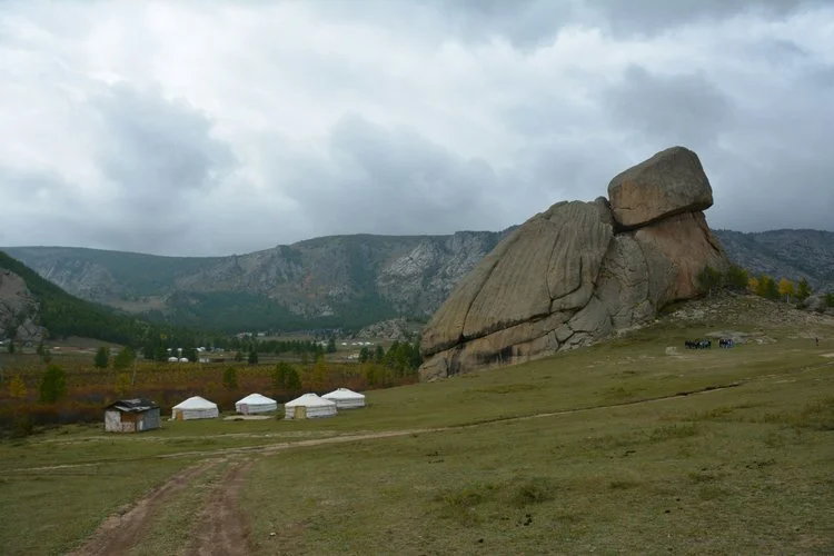 A large boulder resting on smaller rocks on a grassy field, with a mountain range and cloudy sky in the background.