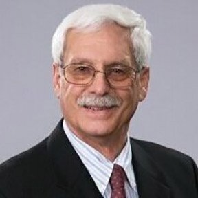 A smiling older man with white hair and glasses, wearing a dark suit, white shirt, and maroon tie, against a gray background.