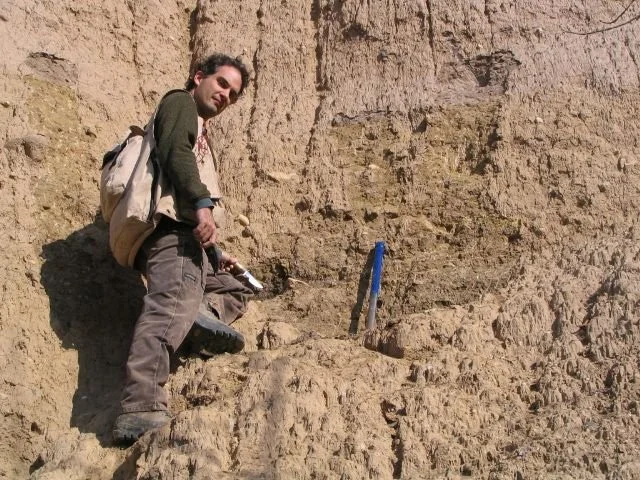 A man standing on rocky terrain holding a rock hammer, with a backpack, near a blue marker in a desert or canyon landscape.