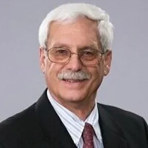 A portrait of an older man with white hair, glasses, and a mustache, wearing a dark suit, white shirt, and a red tie, smiling against a light gray background.