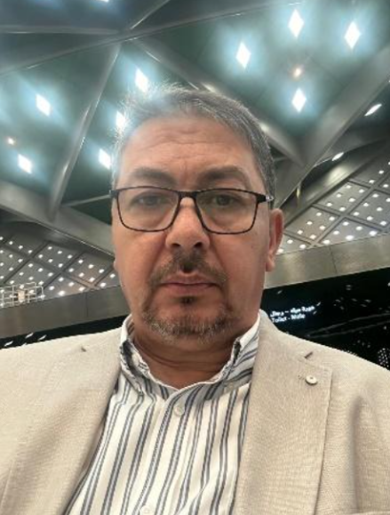 A man wearing glasses and a beige blazer, taking a selfie in an indoor location with a modern, metallic ceiling and bright lights.