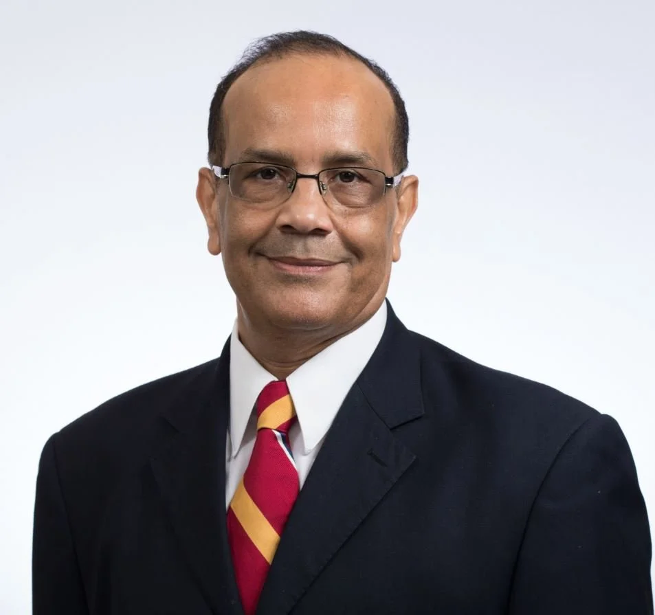 A middle-aged man wearing glasses, a dark suit, white shirt, and a red and yellow striped tie, smiling against a plain white background.