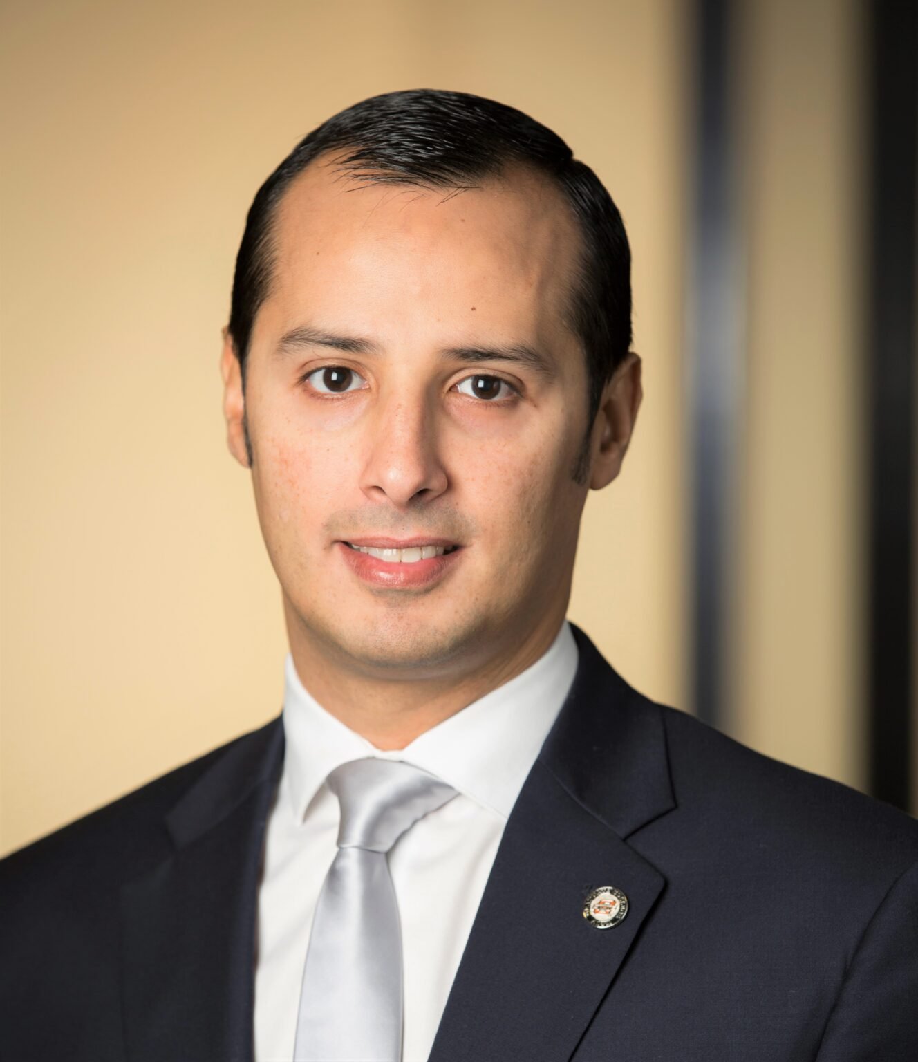 Professional headshot of a man in a dark suit, white shirt, and light-colored tie, with a pin on his lapel, standing against a blurred background.