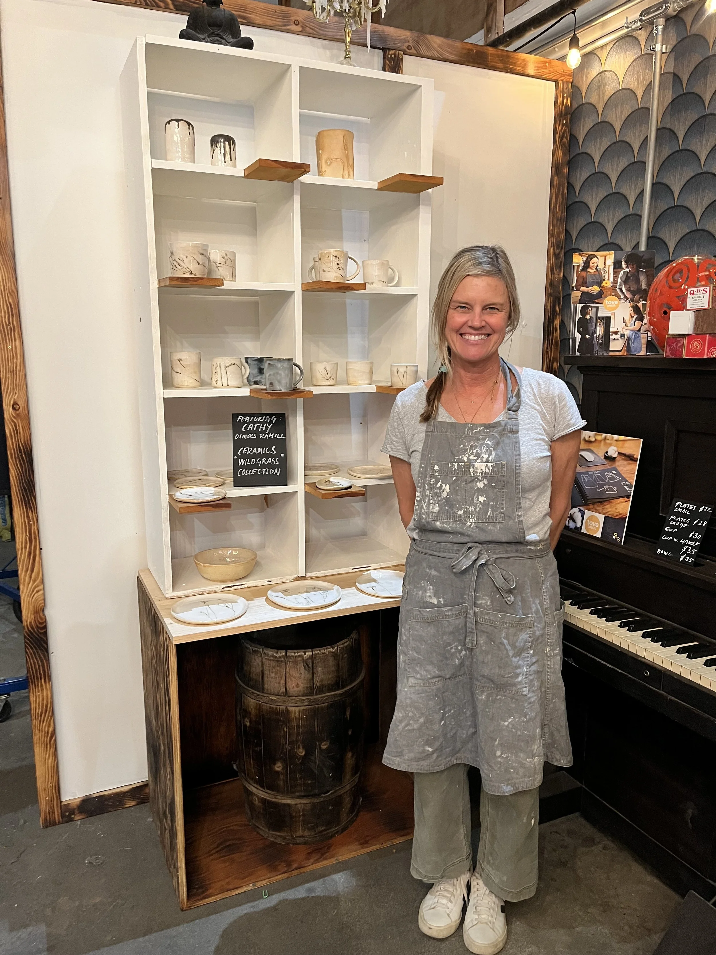 A woman standing in front of a display shelf with ceramic mugs and plates, smiling, wearing an apron and casual clothes in a cafe or art studio setting.