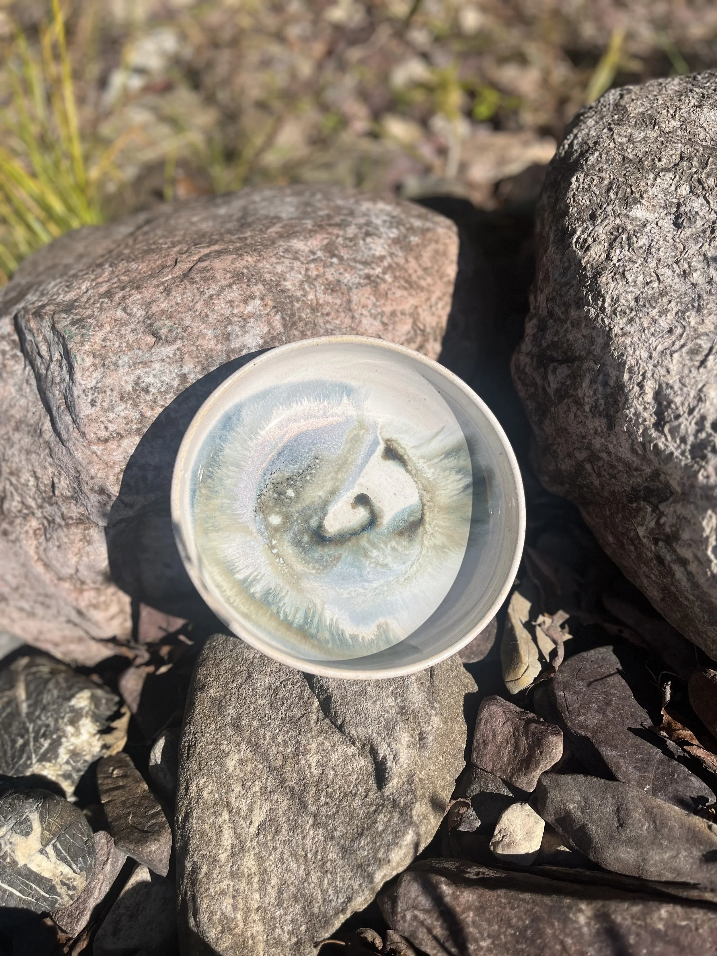 A small round ceramic dish with a swirling, iridescent glaze pattern on a bed of various rocks and stones.