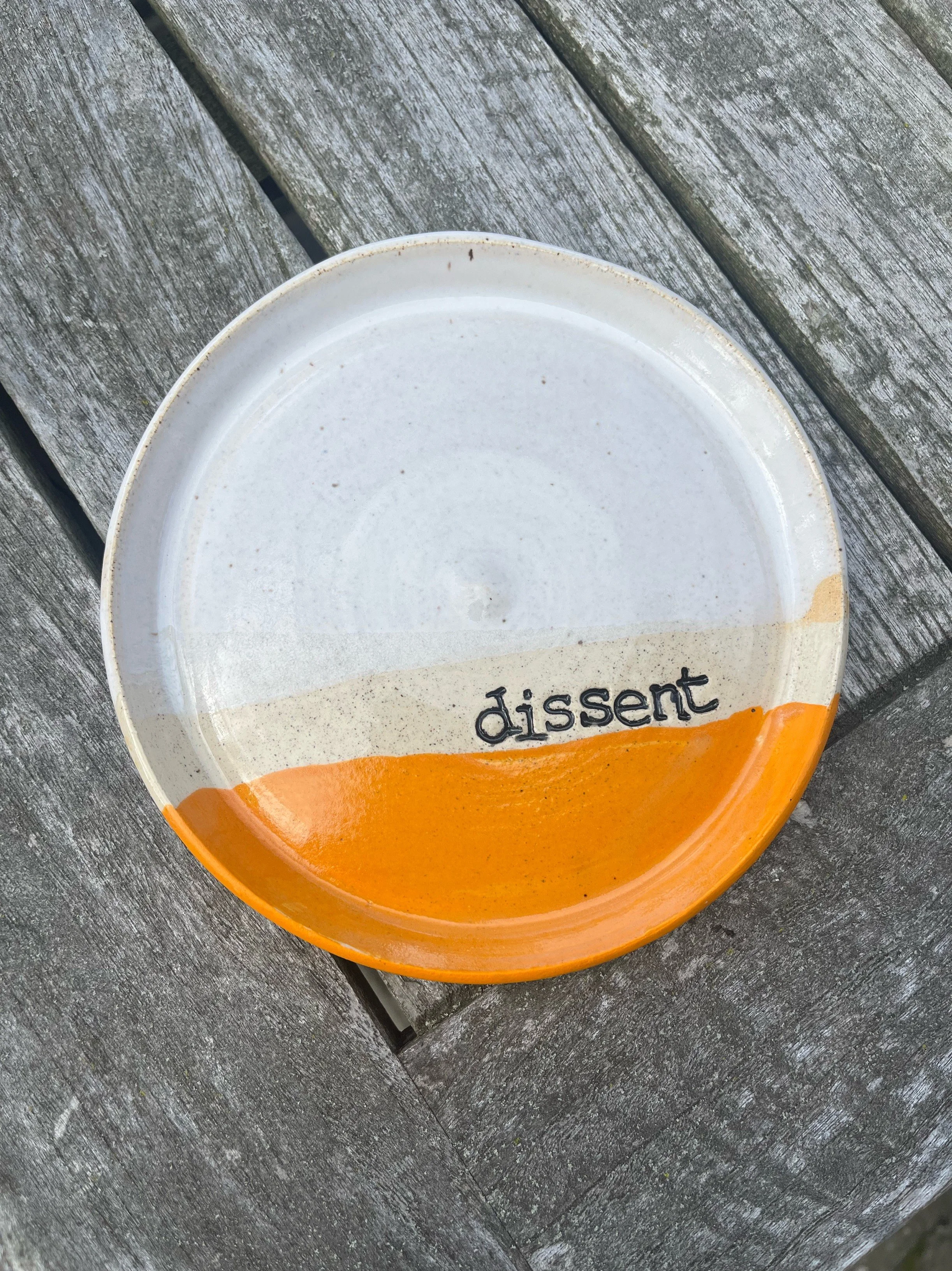 A ceramic plate with a cream background and orange bottom, with the word 'dissent' written on it, placed on a weathered wooden surface.