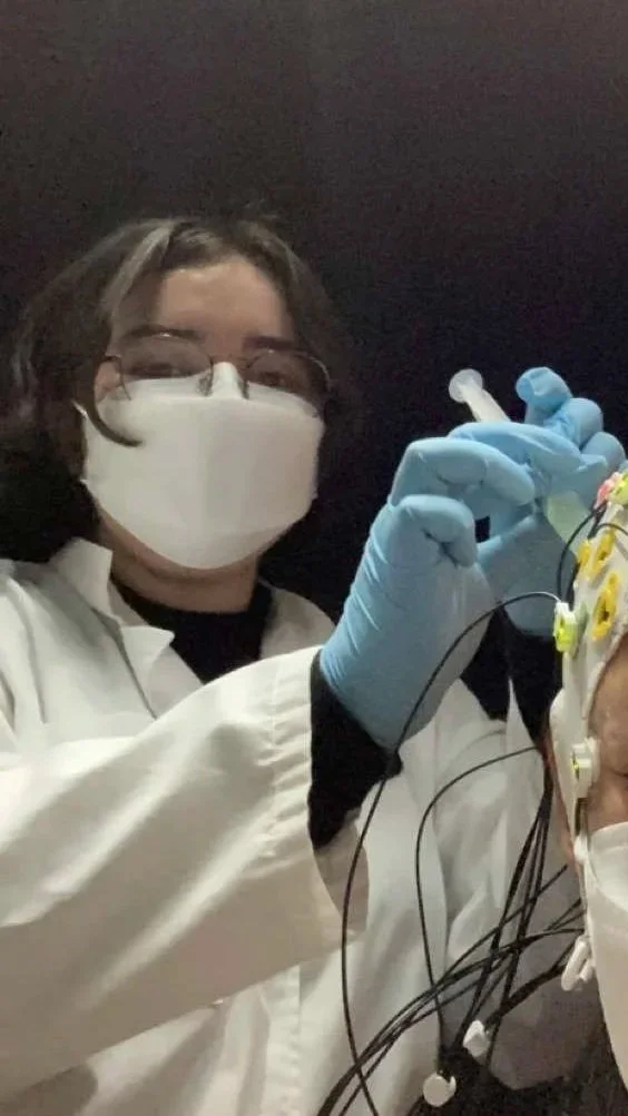 A scientist or medical professional wearing a white lab coat, a mask, and glasses, holding a small vial or syringe, working with wiring and electrodes attached to a person's head in a clinical or laboratory setting.