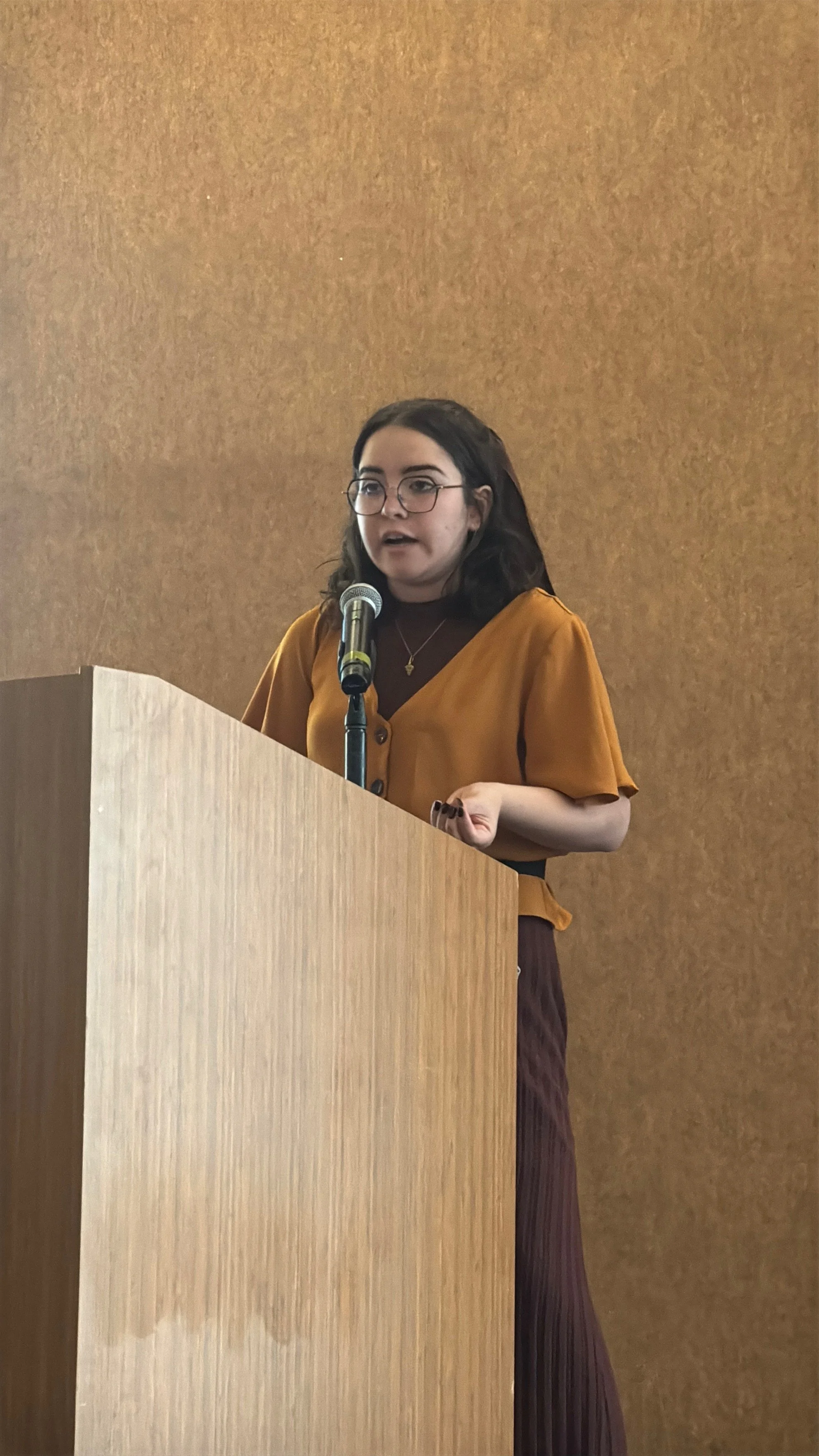 A young woman with glasses is speaking at a podium with a microphone against a brown wall.